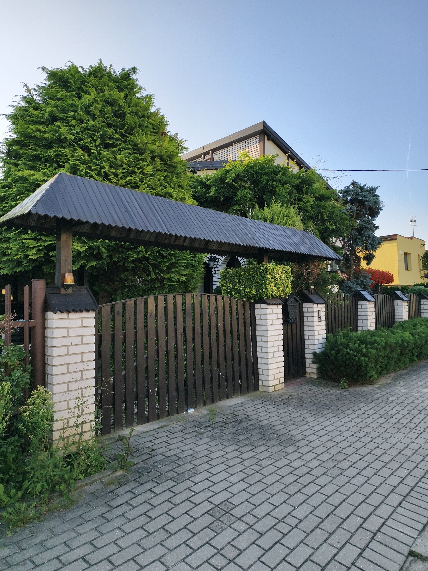 The image shows a segment of a residential property in Tczew, Poland. A dark wooden fence with brick pillars lines the property, with a tiled walkway running alongside it. Above the fence, a large coniferous tree dominates the left side of the frame, while the roofline of a modern house is visible in the upper right, partially obscured by more greenery. A yellow building is also visible in the distance. The sky is clear and blue, indicating daylight. The scene is static and appears to be a typical street view of a suburban area.