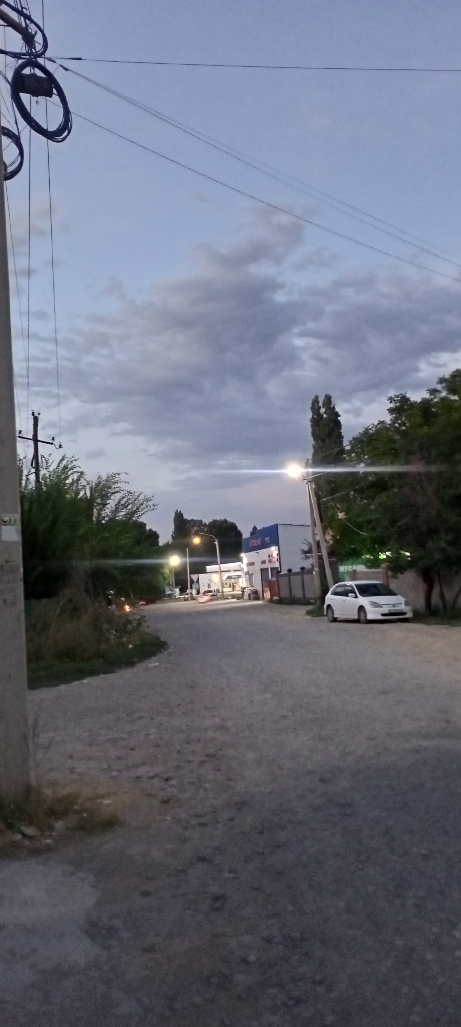 The image depicts a street scene at dusk in Romanovka, Kyrgyzstan. The sky is overcast with heavy clouds, and the light suggests either dawn or dusk. A paved road, appearing somewhat rough and uneven, curves to the left in the foreground and continues towards the background. On the right side of the road, a white sedan is parked. Further down the road, a building complex is visible, featuring a white structure with blue accents and signage. Streetlights are illuminated, casting a warm glow on the surroundings. Lush green trees and bushes line parts of the road and the area around the buildings. On the left side of the frame, a utility pole with power lines and coiled cables is prominent. The overall impression is a quiet, somewhat rural or suburban street on the outskirts of a settlement.
