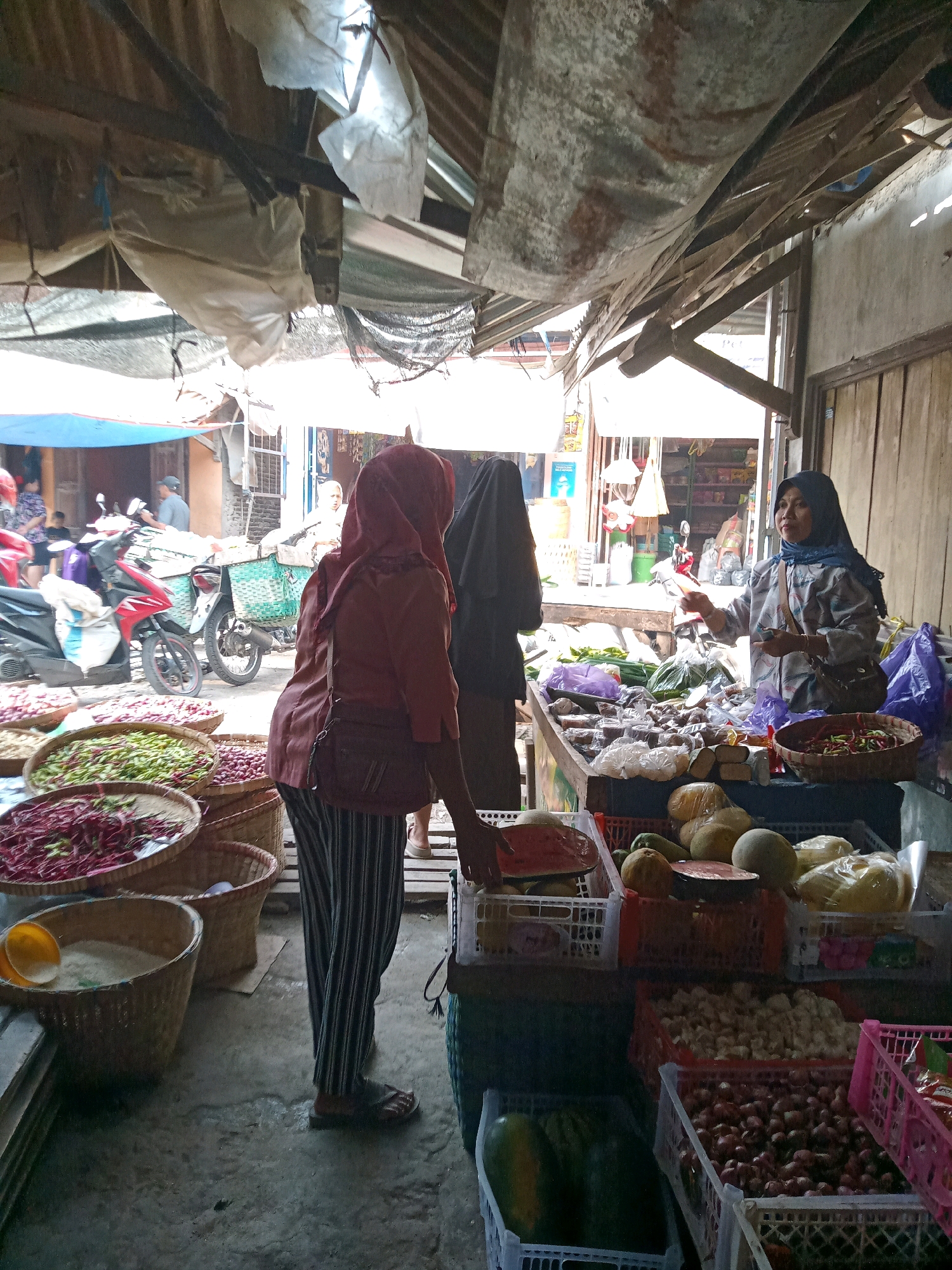 This is a medium shot of an outdoor market in Tanggungharjo, Indonesia. The scene is crowded with various stalls and people. In the foreground, there are several woven baskets filled with produce, including red beans, green beans, and onions. A woman wearing a red headscarf and a patterned skirt is standing next to the baskets, facing away from the camera. To her right, a woman wearing a blue headscarf and a patterned shirt is standing behind a stall, interacting with a customer. The stall is laden with fruits and vegetables, including watermelons, melons, and what appear to be bags of nuts or spices. Behind the stalls, there are motorcycles and other market-goers, as well as awnings and corrugated metal structures providing shade. The overall impression is of a bustling and vibrant local marketplace.