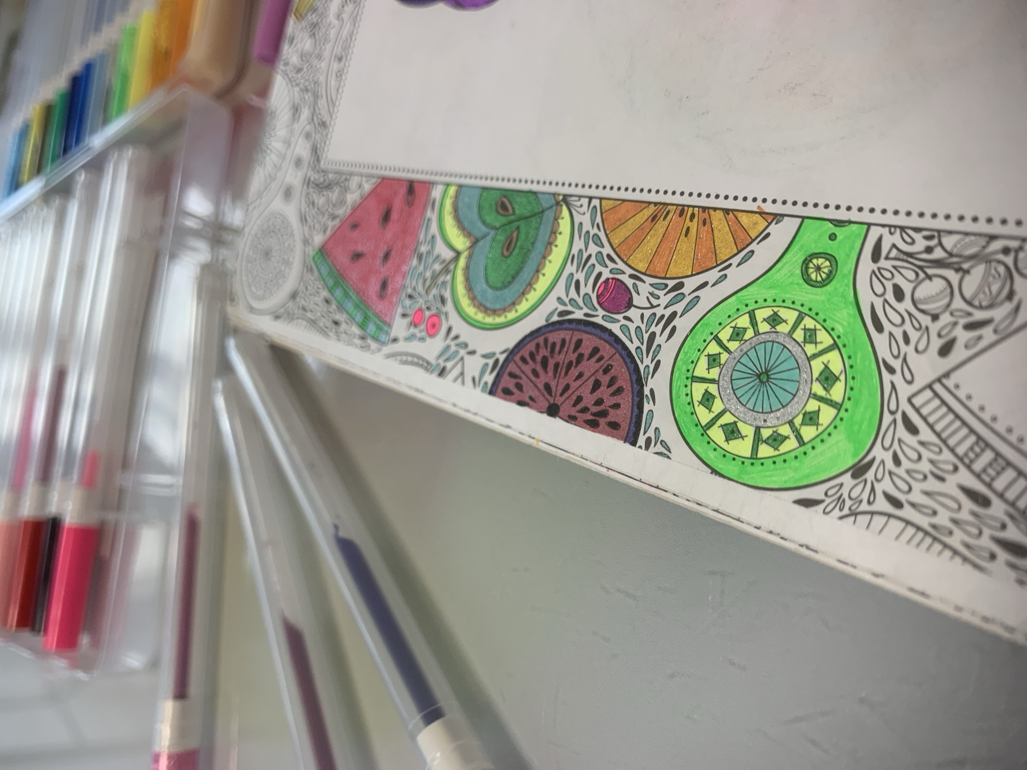 The image presents a close-up, high-angle view of an open coloring book and associated art supplies, located in Izhevsk, Russia. An open coloring book rests on a light gray, textured surface. The left page displays partially colored, intricate designs, including a red watermelon slice with black seeds and a green rind, a heart-shaped green apple slice with a blue interior and brown seeds, a dark red circular fruit slice, an orange fan-shaped fruit slice, and a green circular mandala-like pattern with yellow and silver elements. The right page is largely uncolored, featuring a series of small black dots along its inner margin, indicating a perforated binding. In the blurred background, a transparent plastic container holds numerous gel pens, arranged by color in a spectrum. In the mid-ground, three individual gel pens are visible on the surface: one with a pink cap, one containing purple ink, and a third partially visible. No individuals are present.