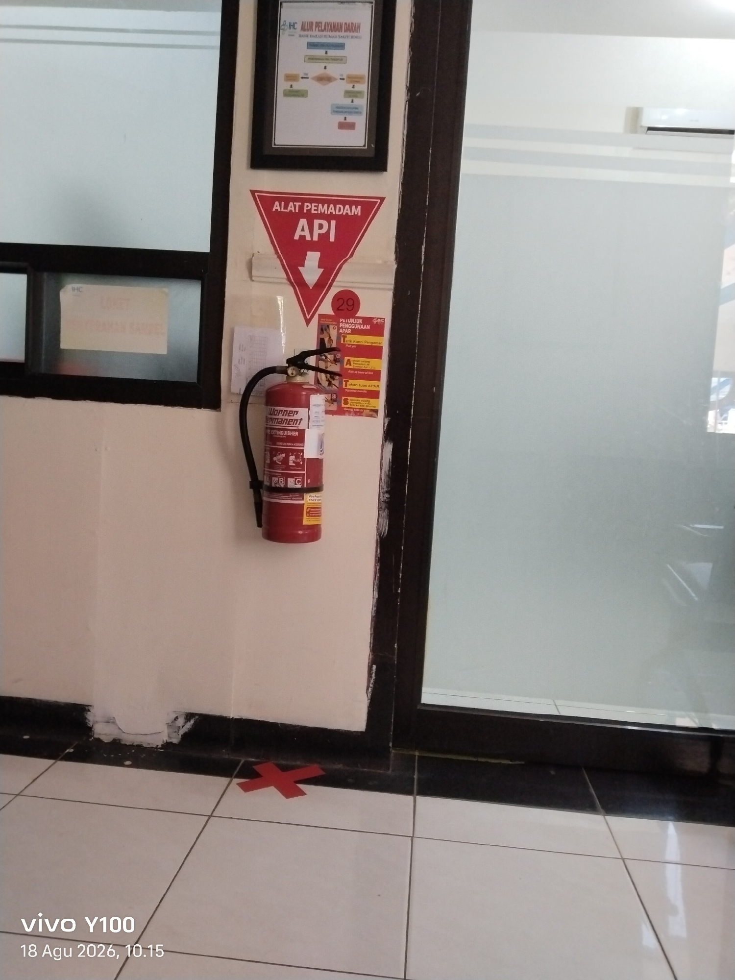 The image depicts an indoor scene with a wall on the left and a reflective glass partition on the right. On the wall, there is a framed poster at the top with text and diagrams, likely an organizational chart or procedure. Below this, a red triangular sign with white lettering reads "ALAT PEMADAM API" which translates to "FIRE EXTINGUISHER API." A red fire extinguisher is mounted on the wall to the right of this sign. Beside the fire extinguisher, there is a yellow instructional poster with the heading "PETUNJUK PENGGUNAAN APAR" (Instructions for using a fire extinguisher). A red "X" marking is visible on the tiled floor in the foreground. The environment appears to be a public indoor space, possibly a hallway or common area, given the presence of signage and floor markings. The lighting suggests an interior environment with artificial or ambient light. The timestamp at the bottom left of the image indicates "vivo Y100 18 Agu 2026, 10.15." The location is stated as Patokan, Indonesia.