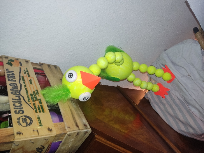 A yellow and green tennis ball bird-like toy is perched on a wooden crate labeled "SICILIANA FRUIT." The toy has a triangular orange beak, googly eyes, a green feather tail, and red cut-out feet. It rests on top of a wooden cabinet next to a white striped cloth. The background consists of a white ceiling and wall.