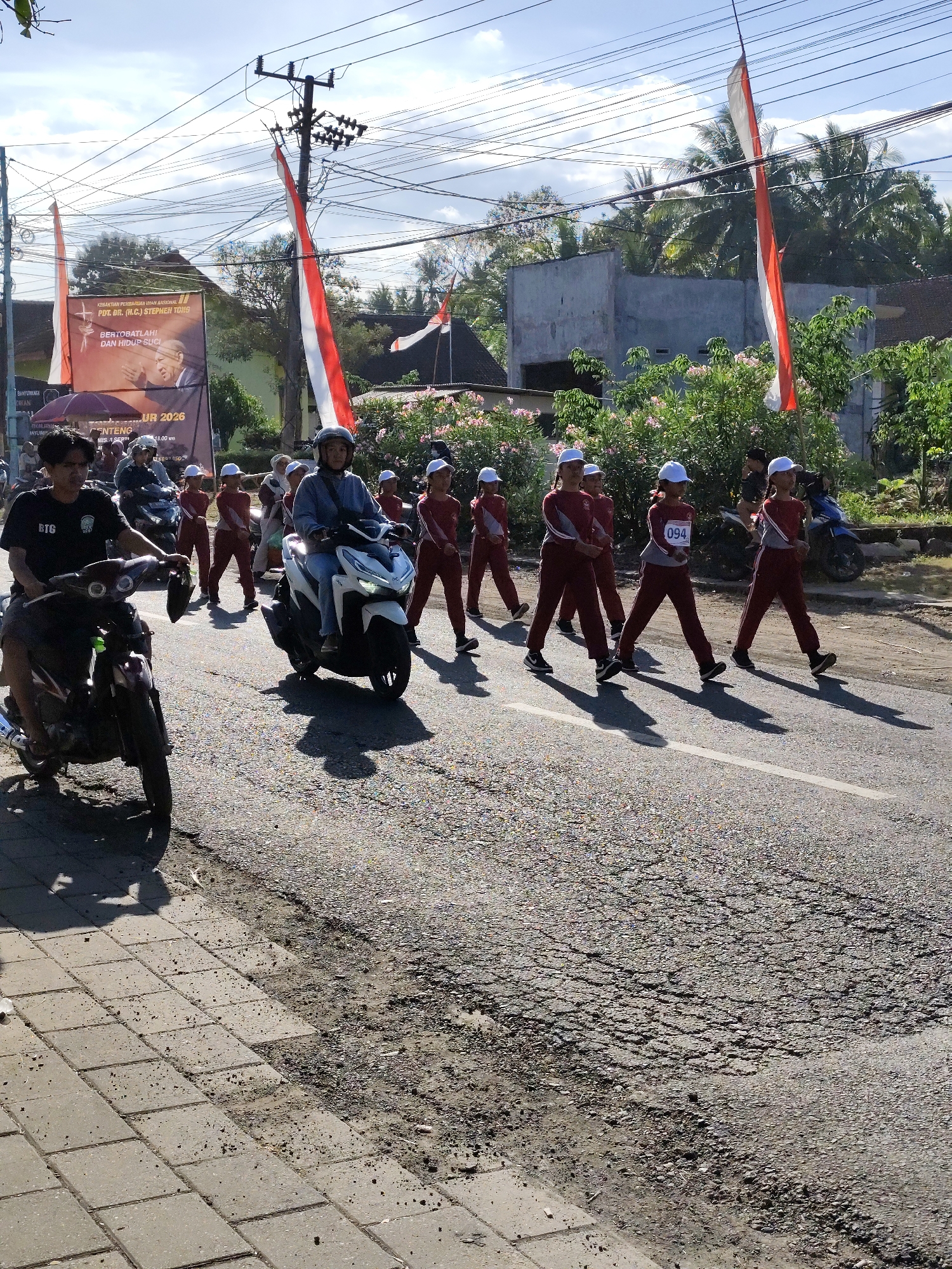 A group of young people, dressed in maroon uniforms and white caps, are marching in formation on a paved road. One of the marchers is wearing a bib with the number "094" on it. They appear to be participating in a parade or procession. Two motorcycles are present in the foreground, one ridden by a person wearing a black shirt and the other by a person in a blue shirt wearing a helmet. In the background, there are buildings, trees, and power lines. Indonesian flags are visible, hanging from poles and attached to a banner. The banner displays text in Indonesian and a portrait of a man. The scene is set outdoors on a sunny day, with shadows cast on the road. The location is identified as Tegalarum, Indonesia.
