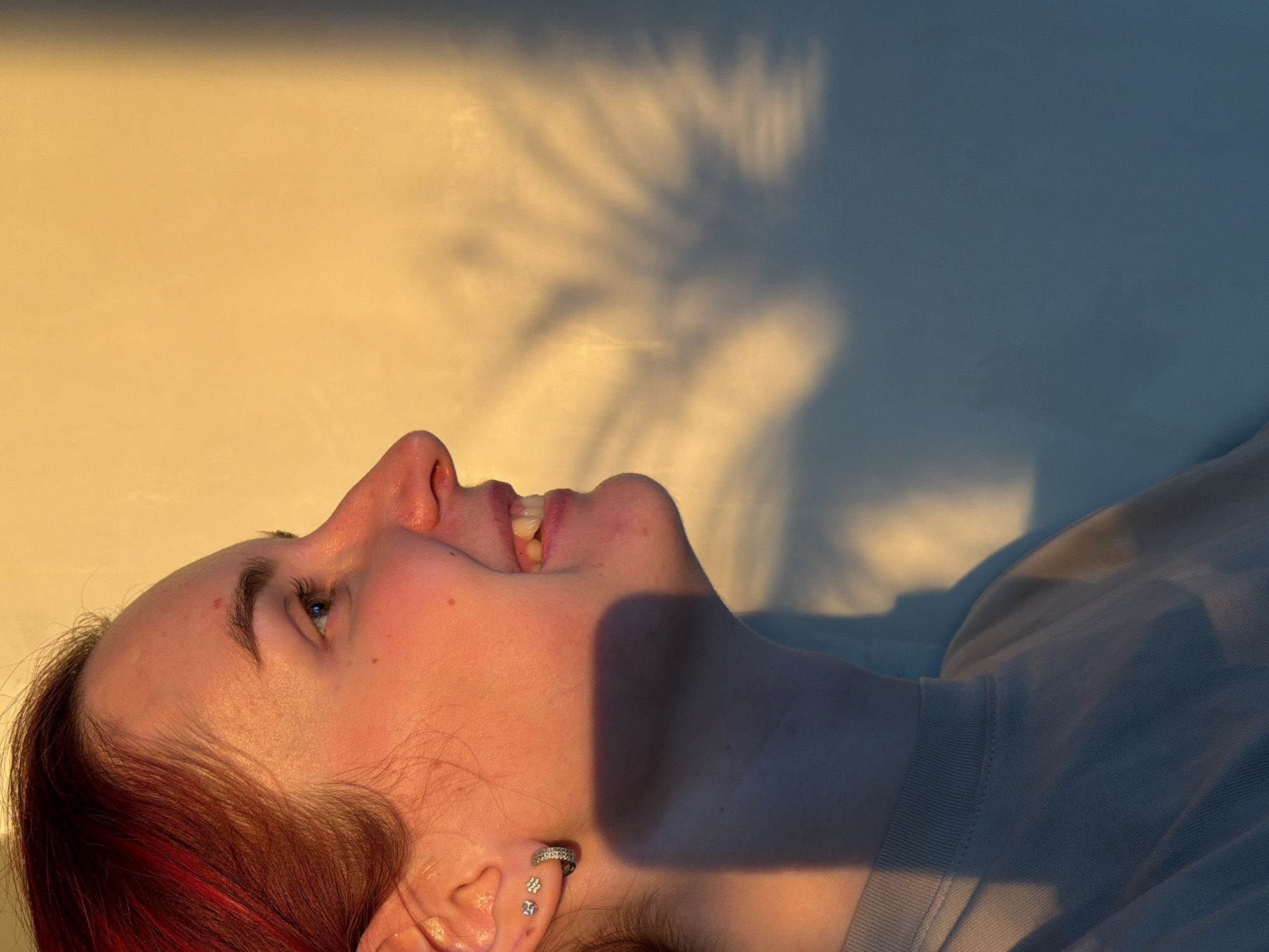 The image displays a young person, likely female, lying down with their head tilted back, looking upwards and smiling or laughing. Their mouth is open, revealing teeth, and their expression conveys joy or amusement. They have fair skin, noticeable freckles on their nose, and red-tinted hair that frames their face. In their visible ear, they wear a silver-colored hoop earring adorned with small stones, and two smaller stud earrings below it. The person is dressed in a light-colored (possibly beige or light grey) t-shirt or sweatshirt.
The setting appears to be an indoor or enclosed space, with a plain wall forming the background. The scene is bathed in warm, golden light, strongly suggesting either early morning or late afternoon, likely sunrise or sunset. This direct, low-angle light illuminates the upper part of the wall and one side of the person's face, creating a strong contrast with the cooler, blueish-grey tones in the shadowed areas of the wall, neck, and clothing. Distinct shadows, resembling tree branches or foliage, are cast upon the wall, hinting at an external light source filtering through a window or opening. The overall mood is warm and positive. No specific location cues for Chaikovsky, Russia, or any visible text are present in the image.