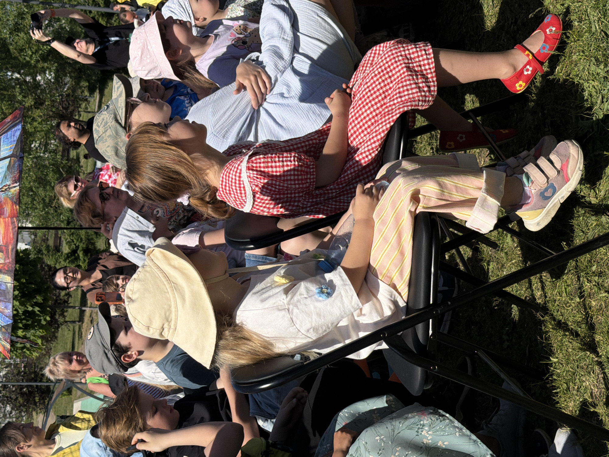 This image depicts an outdoor gathering in the city of Khimki, Russia, on a sunny day. The scene shows a crowd of approximately 15 visible individuals, primarily children and adults, mostly seated on black folding chairs on a grassy area.
In the foreground, two young girls are seated. The girl on the left wears a large cream-colored bucket hat, a loose white t-shirt adorned with blue and yellow crocheted flower appliques, and light pink and yellow striped pants rolled at the cuffs. She has light-colored sneakers with pink and blue accents, and a clear plastic water bottle rests beside her on the chair. The girl on the right has brown hair tied back and is wearing a red and white checkered dress with a white lace collar. She has red Mary Jane-style shoes decorated with colorful flower patterns in red, yellow, and blue. Both girls are looking towards their respective lefts, out of frame.
Behind these girls, several other individuals are visible. To the left, a boy in a dark t-shirt with headphones is partially seen. Further back, another boy wears a camouflage baseball cap, looking to his right. An adult woman with blonde hair and a yellow top is in the upper left. In the midground, a child in a pink bucket hat is visible. Several other adults are scattered, including a woman with dark hair and glasses, and a man with a beard wearing a dark t-shirt. In the upper right background, a man in a black t-shirt holds a camera aloft, seemingly taking a picture. A colorful umbrella with a painted design provides shade further back.
The ground is covered with green grass, illuminated by strong sunlight, casting visible shadows from the chairs and people. Trees and some indistinct structures are present in the distant background, indicating a park or open recreational area.