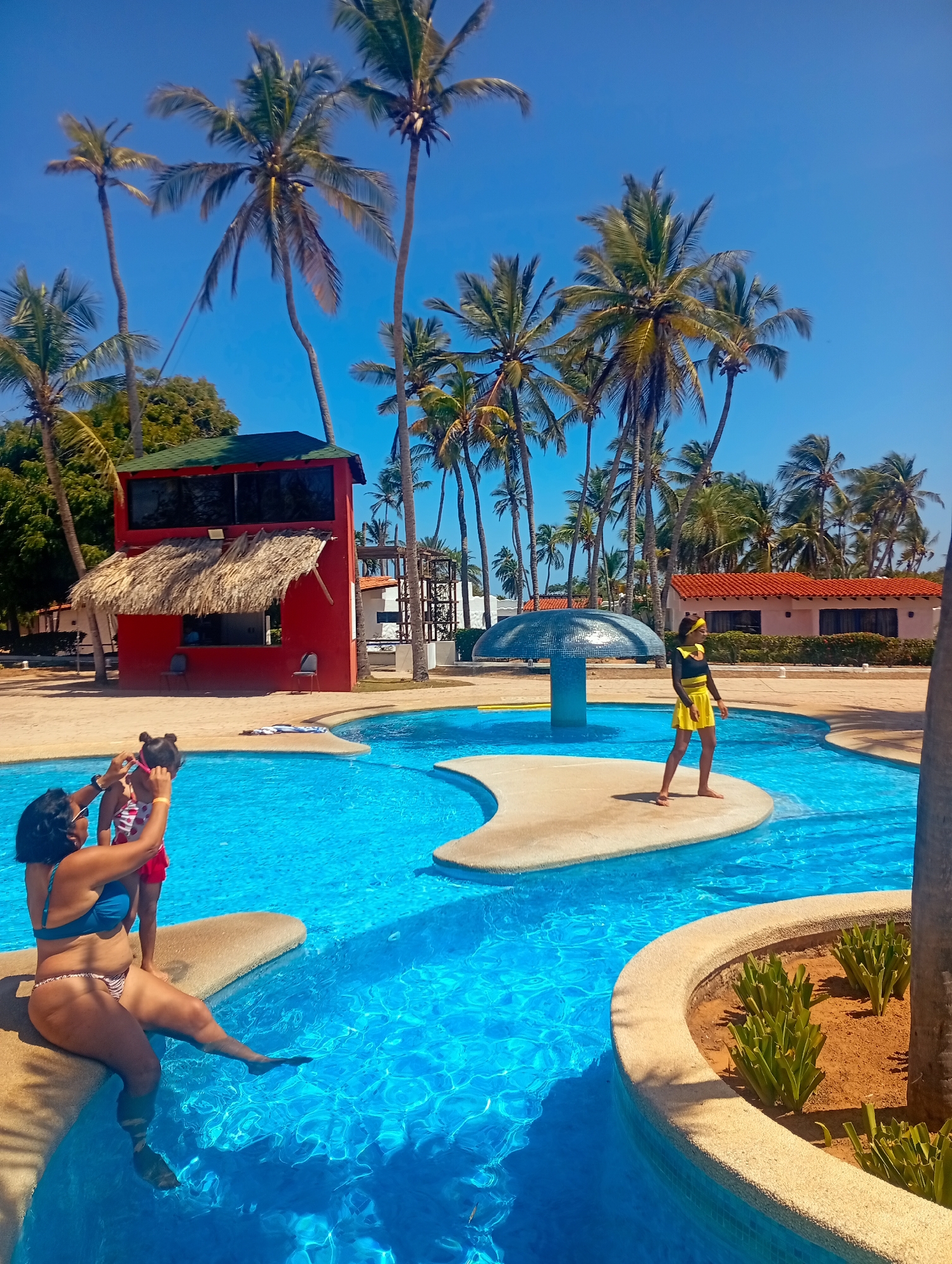 A daytime scene shows a swimming pool complex at Playa El Agua, Venezuela, beneath a clear blue sky dotted with palm trees.
In the foreground, a woman wearing a blue swimsuit sits on the pool's edge, attending to a young child, presumably fitting swimming goggles on the child's face. The pool water is a vibrant turquoise.
In the middle ground, a young woman wearing a black top and a yellow skirt stands on a small, elevated island in the pool. Behind her, a decorative, mushroom-shaped structure is visible. Beyond that, a single-story building with a red-tiled roof stands on the edge of the pool area.
To the left, a two-story red structure with a thatched roof overhang is present. The pool complex appears well-maintained and landscaped with greenery.