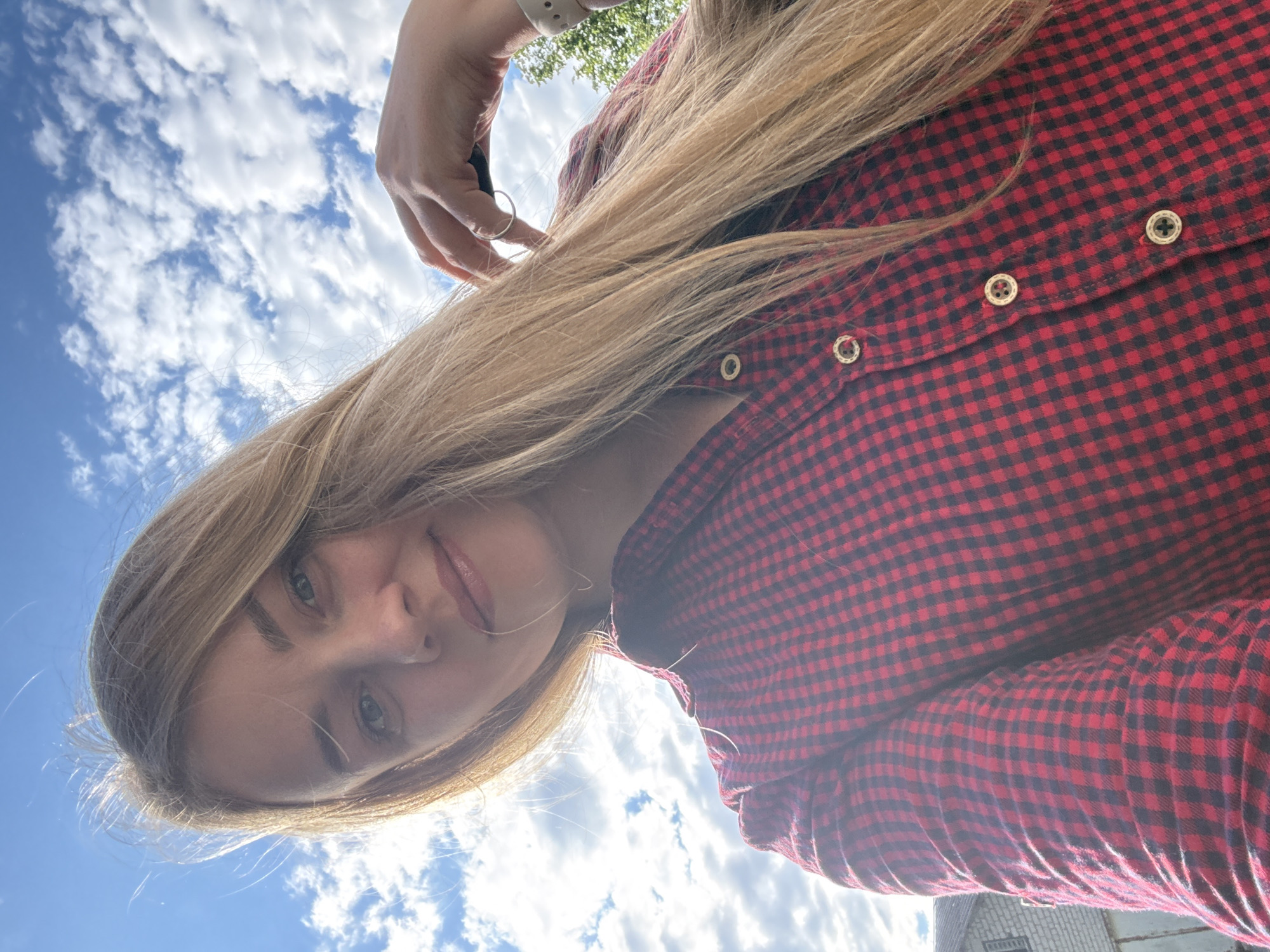 A low-angle, diagonal selfie captures a young woman outdoors in Chaikovsky, Russia. She has long, blonde hair and is wearing a red and black checkered shirt with gold-colored buttons. Her face, including her eyes, nose, and lips with a hint of lip gloss, is visible, and she has a neutral to slightly smiling expression while looking towards the camera. A silver-colored ring is on her finger, and a light-colored wristband is visible on her left wrist, suggesting she is holding the camera.
The background consists primarily of a bright blue sky filled with scattered white, fluffy clouds, indicating a sunny daytime. Green foliage from trees or bushes is visible near the top of the image. A portion of a building, appearing to be constructed from brick or concrete blocks, is visible in the bottom right corner. The bright sunlight creates some glare, particularly near her face and the sky on the right side. There is no visible text in the image.