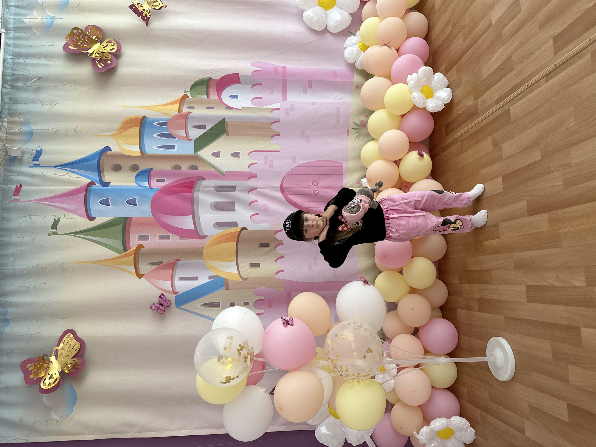 A young girl lies on the floor in front of a backdrop decorated with a cartoon castle and butterfly cutouts. The backdrop appears to be part of a celebratory or decorative arrangement, possibly for a birthday or other special occasion. The girl is wearing a black shirt, pink pants with a cartoon character on them, white socks, and a black baseball cap. She holds a stuffed animal in her arms. The floor is made of light brown wooden planks. An arrangement of pastel-colored balloons, interspersed with artificial white flowers, frames the backdrop. The location is Chaikovsky, Russia.