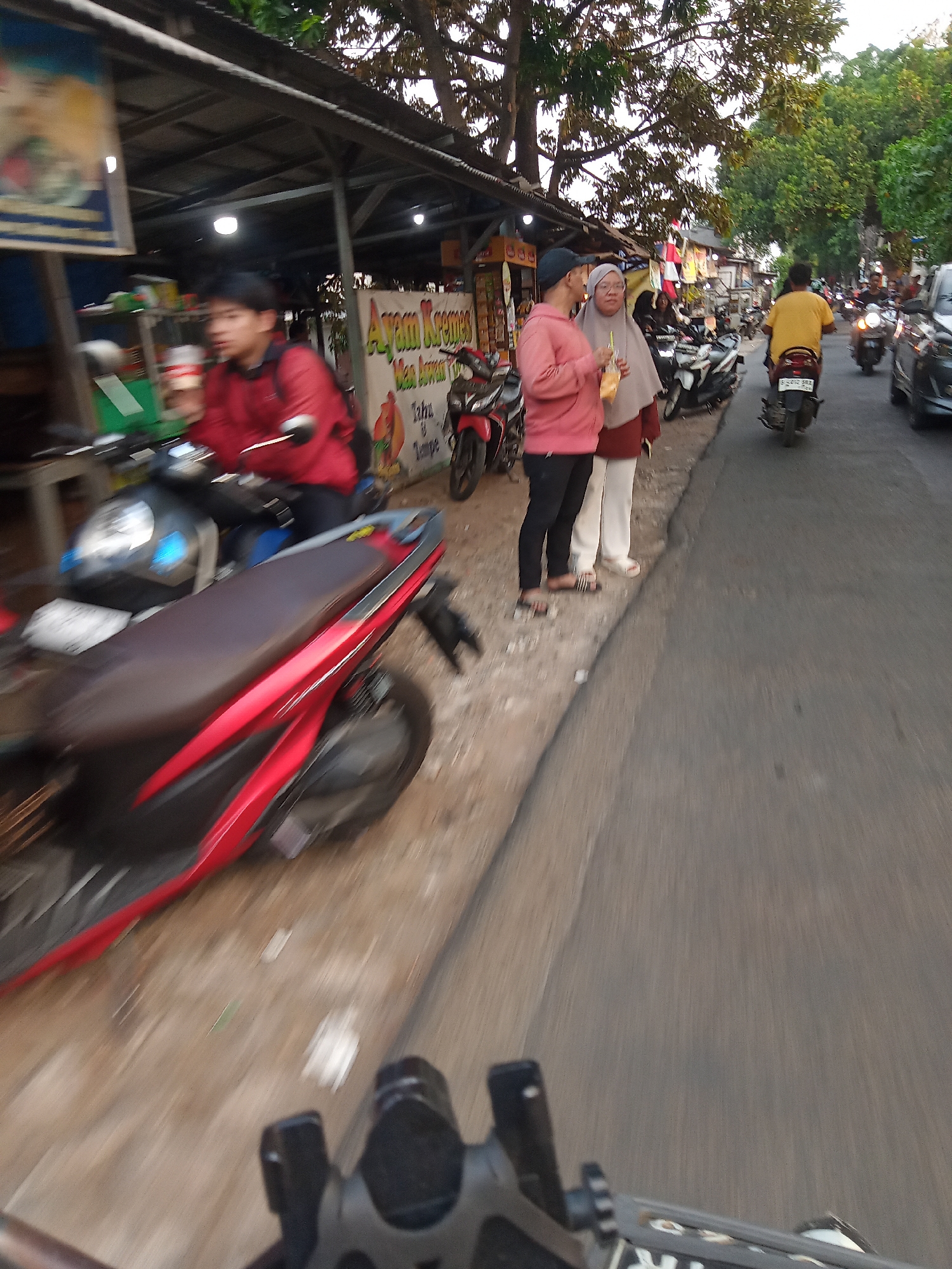 The image captures a street scene in South Tangerang, Indonesia, possibly in a commercial or residential area with roadside vendors. In the foreground, a red and black scooter is partially visible, suggesting a first-person perspective, likely from someone riding or standing near it. The background reveals a bustling street with several individuals and vehicles.
On the left, a young man wearing a red jacket is on a dark-colored scooter, holding a drink. Beside him, a stall with a metal roof displays a sign with Indonesian text, likely advertising food. Further down the street, two women are standing on the sidewalk. One, wearing a pink hoodie and black pants, holds a drink with a straw. The other, in a beige headscarf and white pants, stands next to her.
The street is lined with various stalls and shops, suggesting a local market or business strip. Several other motorcycles are parked or moving on the street, indicating typical traffic in the area. A person in a yellow shirt is riding a motorcycle further down the road, and other vehicles, including cars, are visible. The overall environment appears to be an urban setting with moderate activity, characteristic of South Tangerang. The lighting suggests it is either early morning or late afternoon.