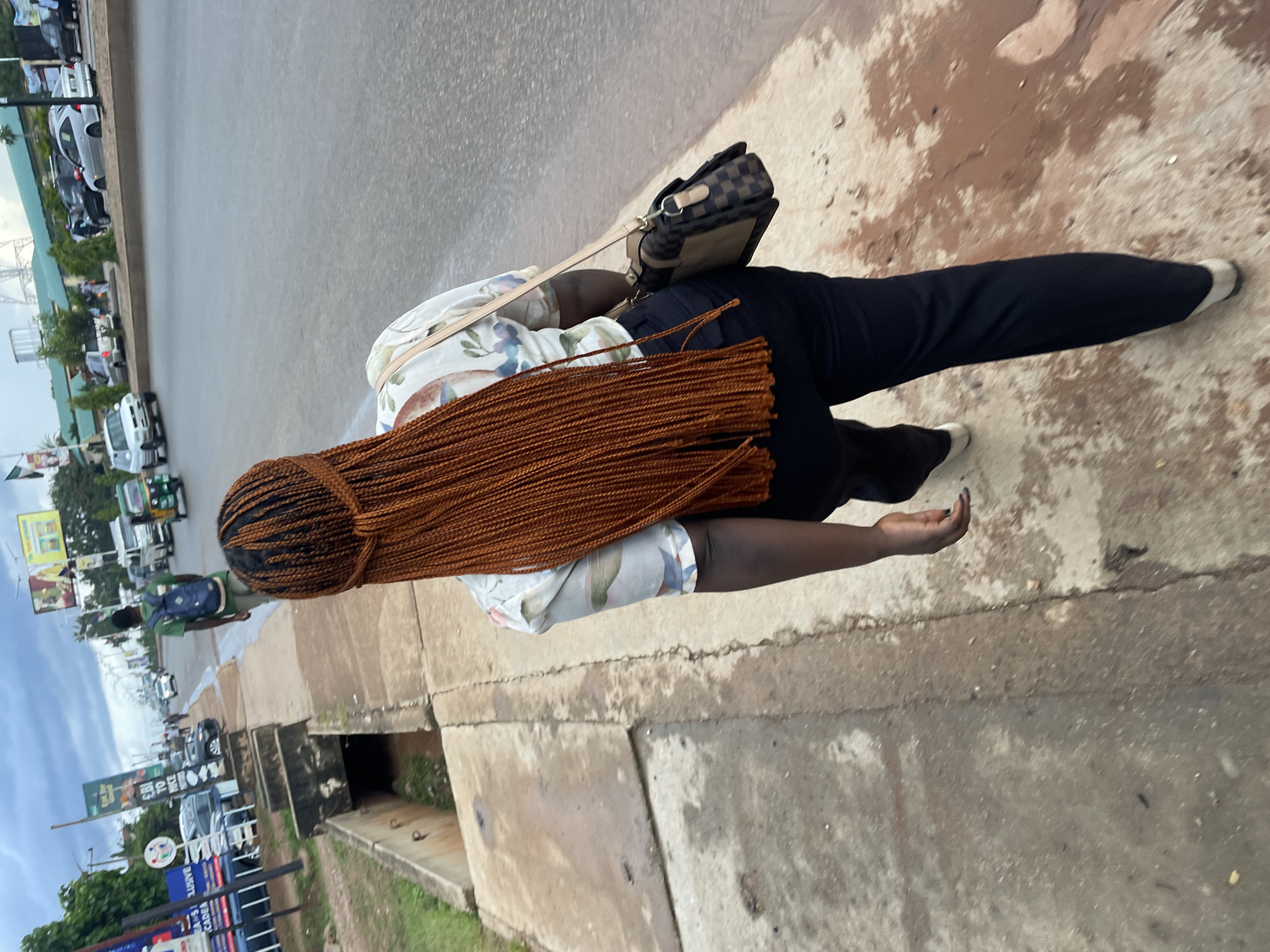A woman with long, braided, copper-colored hair is walking away from the camera on a paved sidewalk next to a street. She is wearing a light-colored shirt with a floral pattern and dark pants. A handbag with a checkered pattern is slung over her shoulder. The street is busy with traffic, including cars, a tuk-tuk, and a motorcycle. In the background, there are buildings, billboards, and other pedestrians. The scene appears to be in Jos, Nigeria, based on the presence of distinct billboards and architectural styles common to the region. The sky is overcast, suggesting it might be a cloudy day.