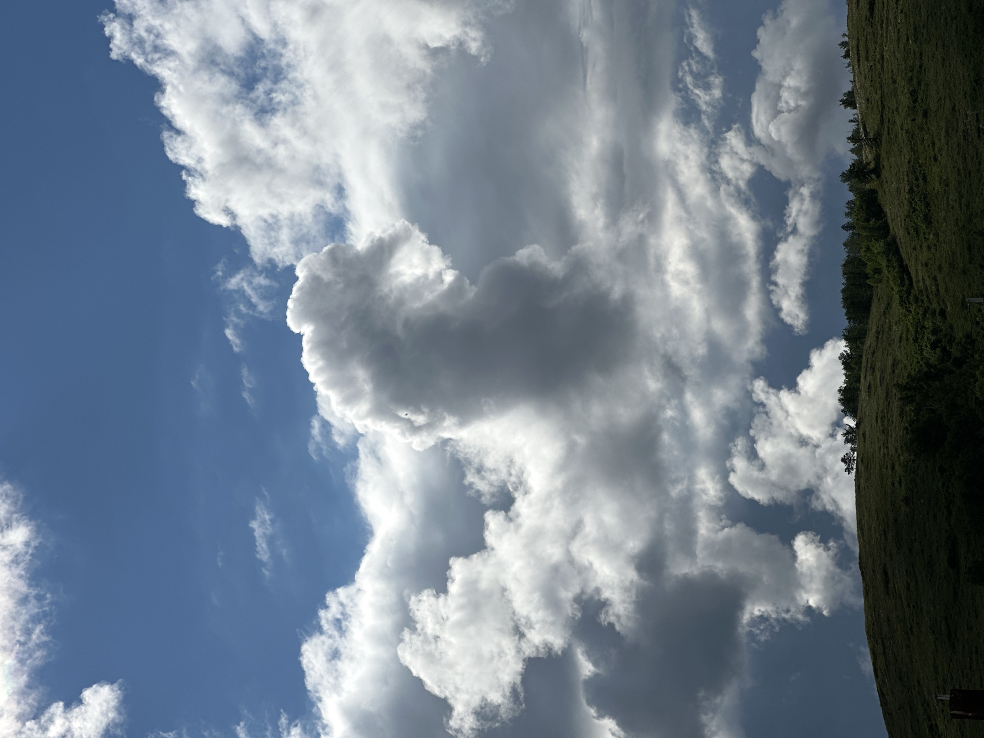 The image shows a daytime scene with a view of the sky. Dominating the scene are large, billowy clouds, ranging in color from bright white to dark gray, suggesting varying thicknesses and densities. Patches of blue sky are visible between the clouds, indicating a partly cloudy day. To the right of the image, there is a section of land covered in green vegetation, possibly a hillside or field. The image appears to be rotated or taken at an angle. There is no visible activity related to people or interactions between people and objects. The location appears to be near a green, grassy hillside under partly cloudy skies.