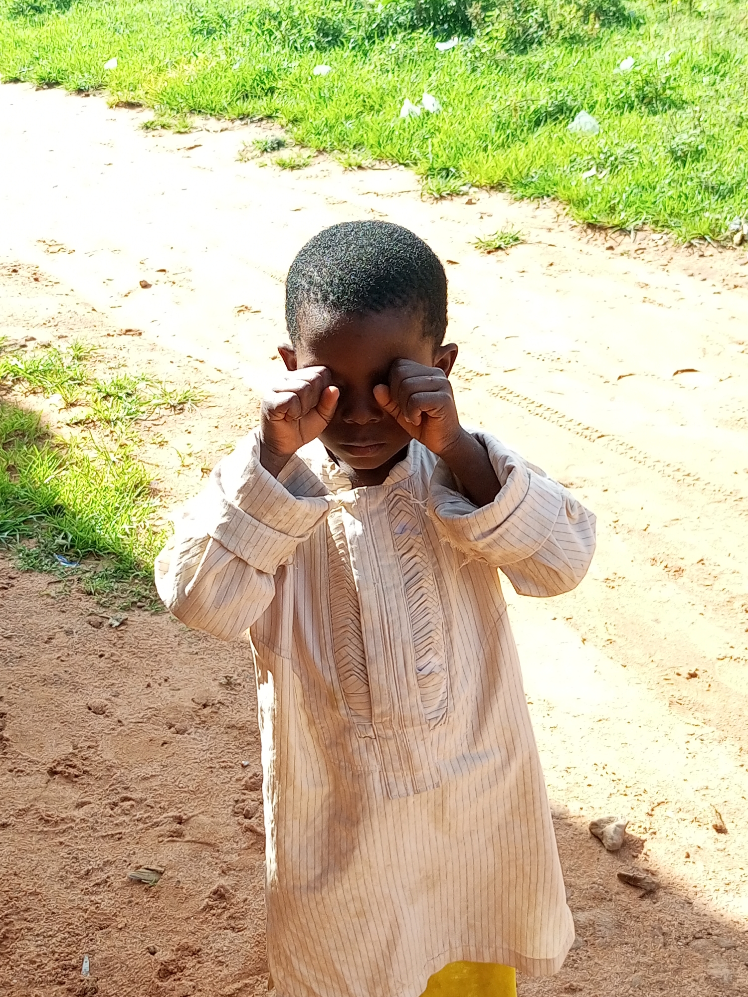 A young child, appearing to be of African descent, stands in what seems to be a rural or undeveloped area, possibly on the outskirts of Tempure, Nigeria. The child is wearing a light-colored, striped tunic and has their hands covering their eyes, suggesting they might be crying or rubbing their eyes. The ground is a mixture of sand and dirt, with tire tracks visible in the background, indicating it may be a pathway or road. Lush green grass lines the left side of the image, with some white debris scattered within it. The lighting suggests it is daytime, with the sun casting shadows.