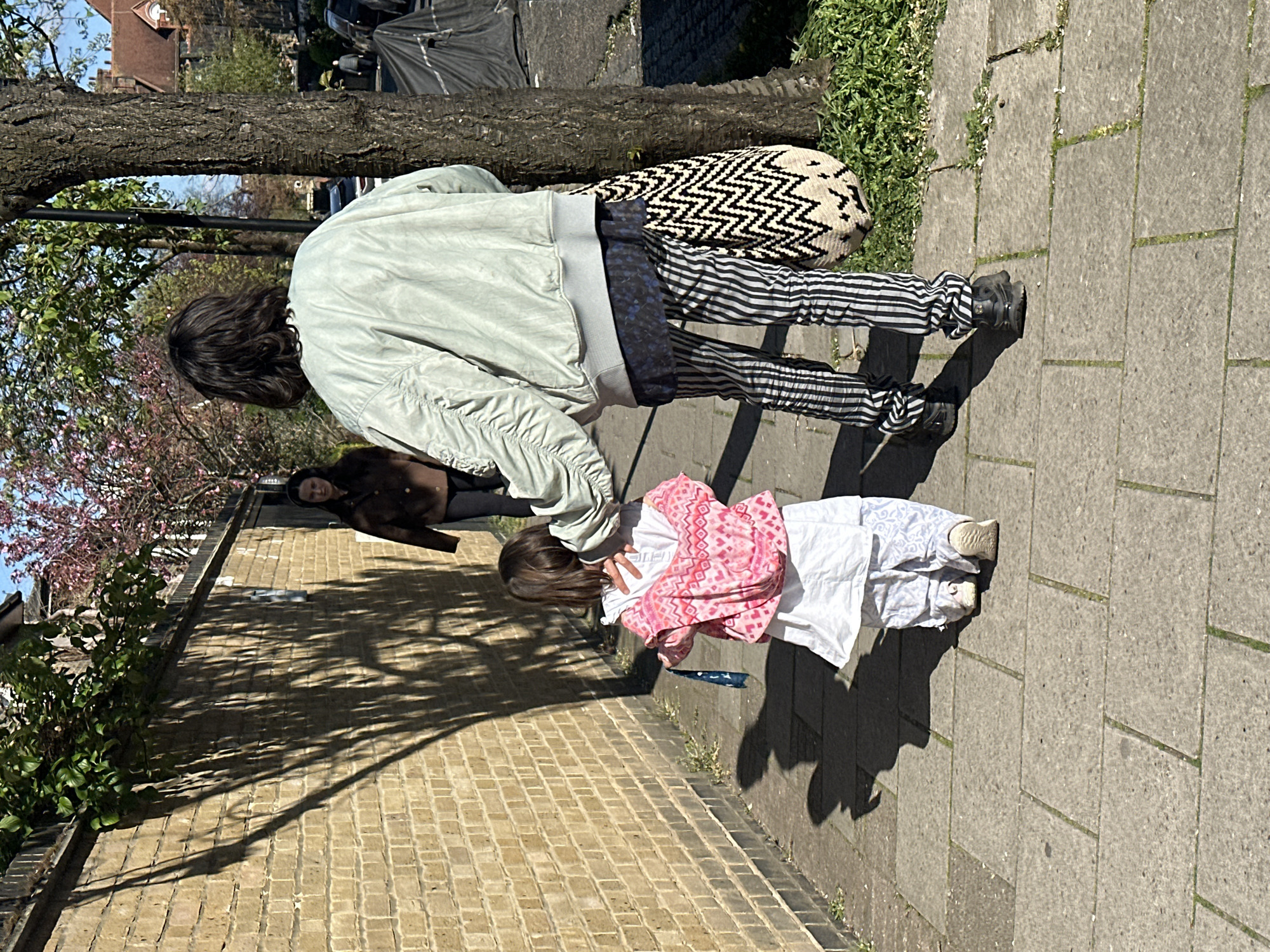 The image depicts an outdoor urban scene, likely in London, United Kingdom, suggested by the architecture and street features. A person wearing a light green jacket, black and white striped pants, and black shoes is standing next to a young child who is wearing a pink patterned top and white pants. The person appears to be interacting with the child, possibly adjusting their clothing or offering assistance. The child is facing the person, seemingly engaged in the interaction.
Behind them, a tree trunk is visible, and further in the background, another person dressed in brown is walking in the opposite direction. The setting includes a brick wall on one side and a paved sidewalk. The scene is illuminated by bright daylight, casting shadows of the individuals and objects present.
Cars parked on the side of the street and residential buildings are present, adding to the typical urban environment. The presence of flowering trees suggests it is likely springtime.