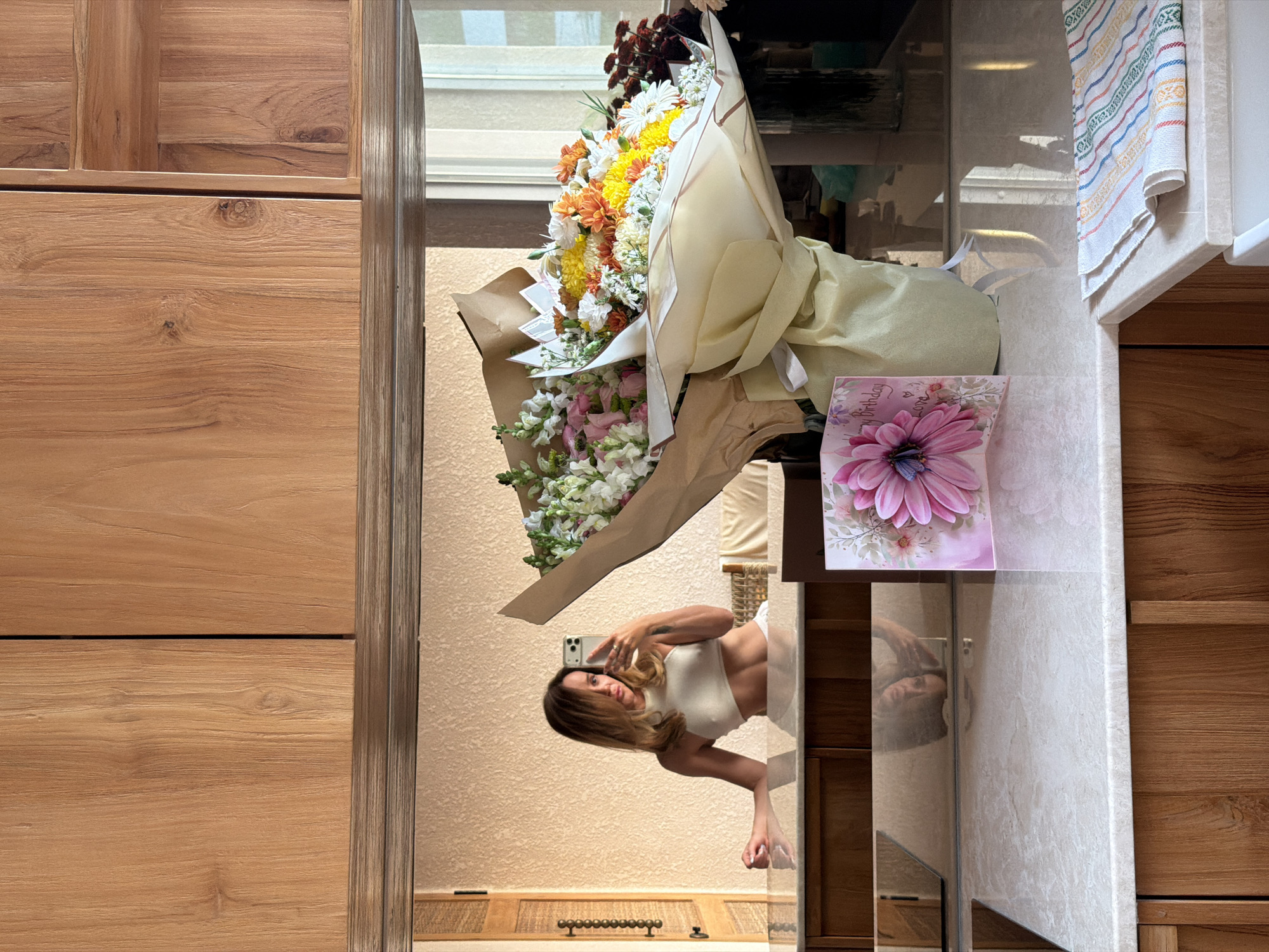 A bathroom mirror reflects a woman holding a phone taking a selfie, with a large bouquet of flowers and a birthday card placed on the vanity. The bouquet features various colors, including white, yellow, and orange. The bathroom has wooden cabinets and a patterned towel. The location appears to be inside a residence, but the exact location in Mengwi, Indonesia, cannot be determined from the image alone.