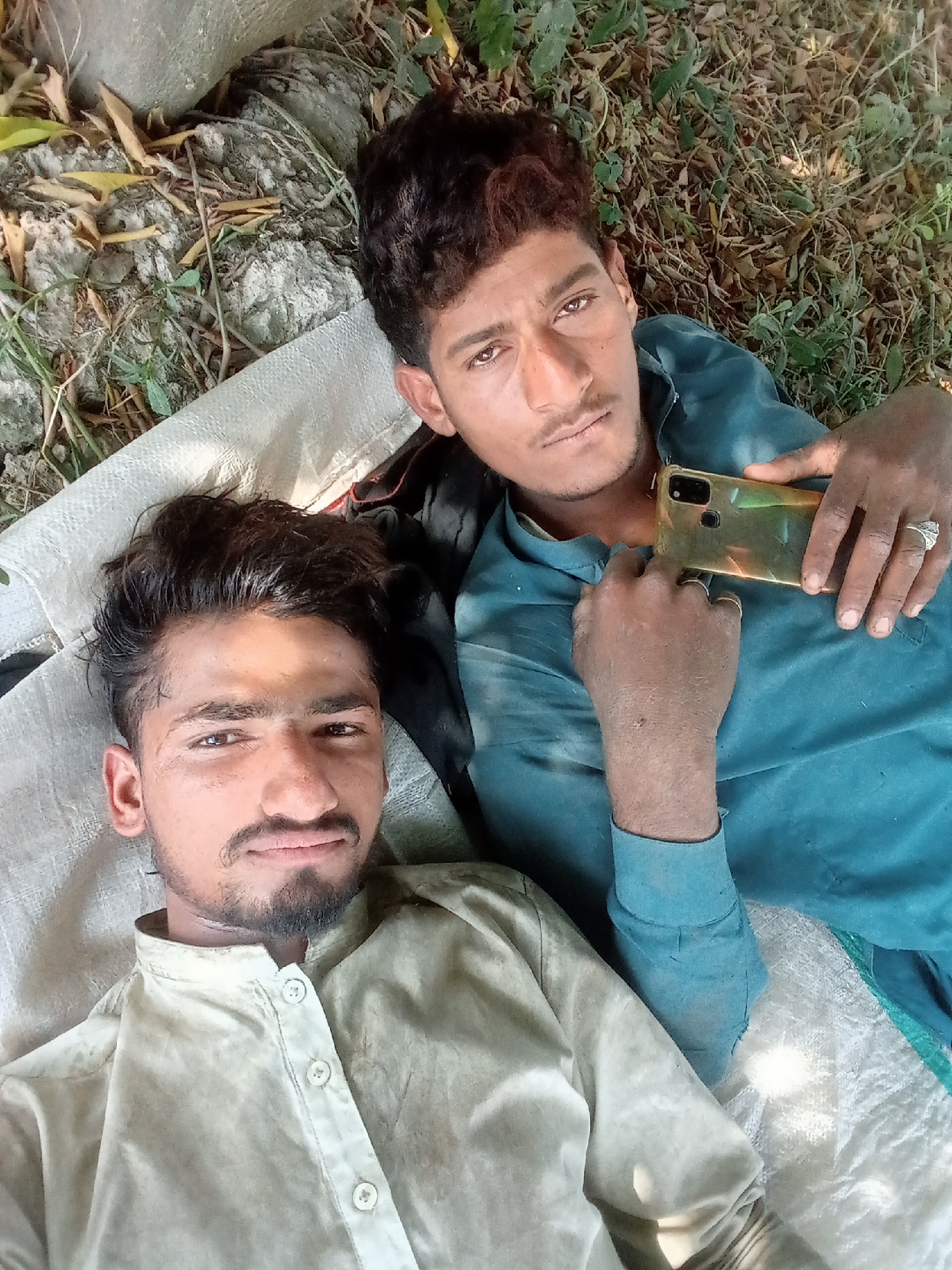 Two men are lying on a white sack or sheet outdoors. The man in the foreground is wearing a light-colored, long-sleeved shirt with buttons. The man behind him is wearing a blue, long-sleeved shirt and holding a gold-colored smartphone. They are outdoors, with some dry leaves and grass visible around them. No specific location in Toba Tek Singh can be determined from this image.