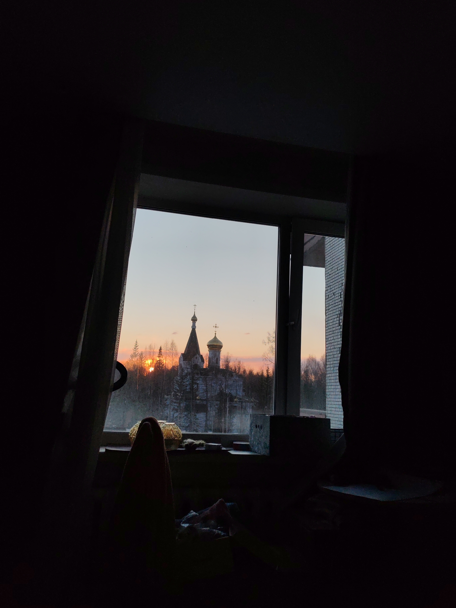 The image, taken from within a darkened room, shows a view through a window. The window is framed by dark curtains, and the interior of the room is indistinct due to low lighting. Outside, the scene reveals a landscape with a church silhouetted against a sunset or sunrise sky. The church features a central spire and at least one gold dome, suggesting Russian Orthodox architecture. Trees surround the church, and snow covers the ground, indicating a winter setting. The location is Ust-Ilimsk, Russia. A light-colored building with a tiled exterior is visible in the background. The windowsill contains various indistinct objects, including a decorative sphere and box.