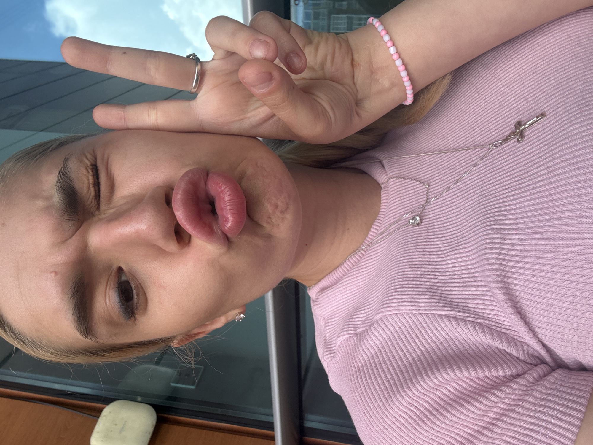 A close-up, rotated selfie shows a young person with light-colored hair, posing playfully outdoors or near a window in Sofia, Bulgaria, under a bright, clear blue sky with some white clouds. The subject is seen winking with their left eye and puckering their lips in a "fish face" expression. Their left hand is raised near their face, forming a peace sign, with a silver-toned ring on the index finger. On their right wrist, a bracelet made of pink and white beads is visible.
The person is wearing a light pink, ribbed short-sleeved top. Adorning their neck is a silver-toned necklace featuring a distinctive cross pendant, which appears somewhat stylized or sword-like, along with a smaller, sparkly pendant. A small, possibly diamond or cubic zirconia, stud earring is visible in their left earlobe. The background elements include a dark-colored building or structure, reflections on glass, and a wooden surface on which a light-colored, rectangular object (resembling an eraser) rests. The image captures a lighthearted moment during daylight hours. No visible text is present.