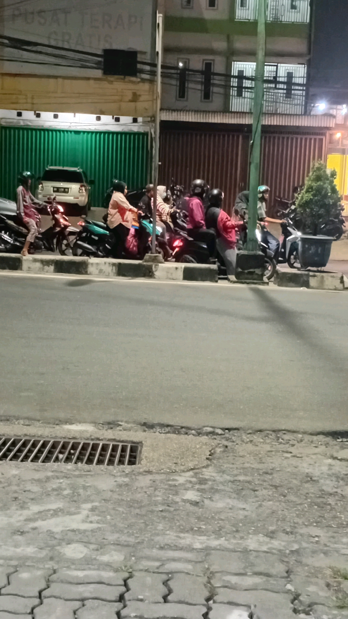 The image depicts a street scene at night in Jambi, Indonesia, focusing on a group of people on motorcycles. In the foreground, a grey asphalt road is visible, with a metal grate for drainage on the left side and patterned paving stones further left. In the mid-ground, a row of motorcycles is parked along a curb. Several individuals are seated on the motorcycles, some wearing helmets and headscarves. One person in a pink jacket is standing near a motorcycle. Behind the motorcycles, a white SUV is parked in front of a green corrugated metal wall. Above this, a building with multiple windows and bars is visible, along with a sign in Indonesian that appears to say "PUSAT TERAPI GRATIS" (Free Therapy Center). The overall impression is a common street scene at dusk or night, possibly near a commercial or service establishment.