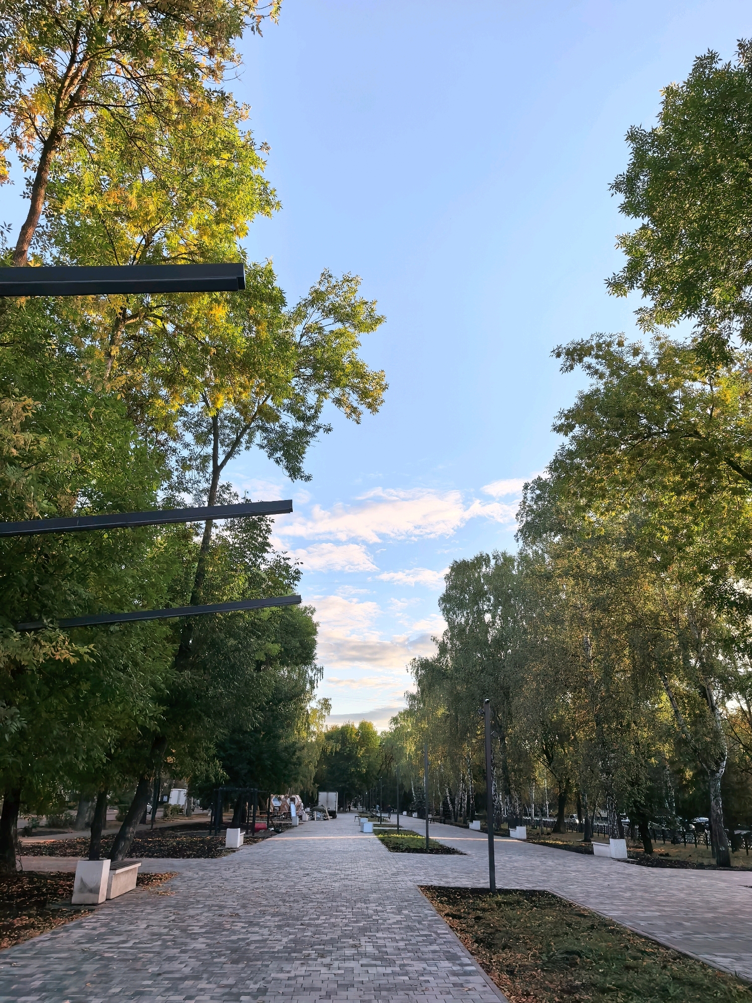 The image depicts a paved pedestrian walkway in a park-like setting, likely in the city of Sterlitamak, Russia. The walkway is lined with trees on both sides, some of which show autumnal foliage in shades of green and yellow. Several modern streetlights and park benches are visible along the path. In the background, there are signs of urban infrastructure, including what appear to be parked vehicles and structures that could be part of a market or fairground. The sky is blue with scattered clouds, suggesting fair weather. The overall scene conveys a sense of a public recreational space.
