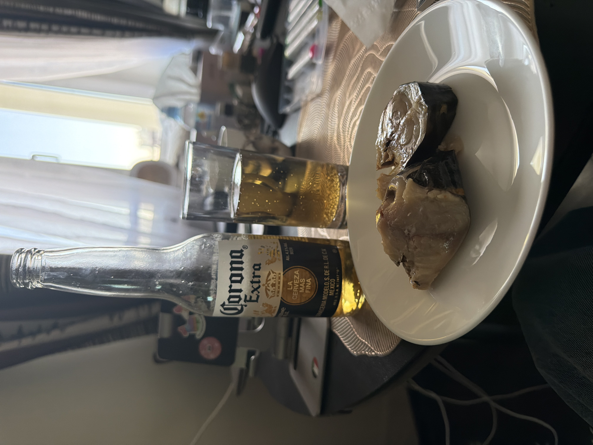 The image shows a Corona Extra beer bottle and a glass of beer next to a white plate with what appears to be pieces of cooked fish. The setting is an indoor environment with a patterned placemat underneath the items. The overall scene suggests a meal or snack. It is not possible to determine the specific location or any action or interaction with the information available.