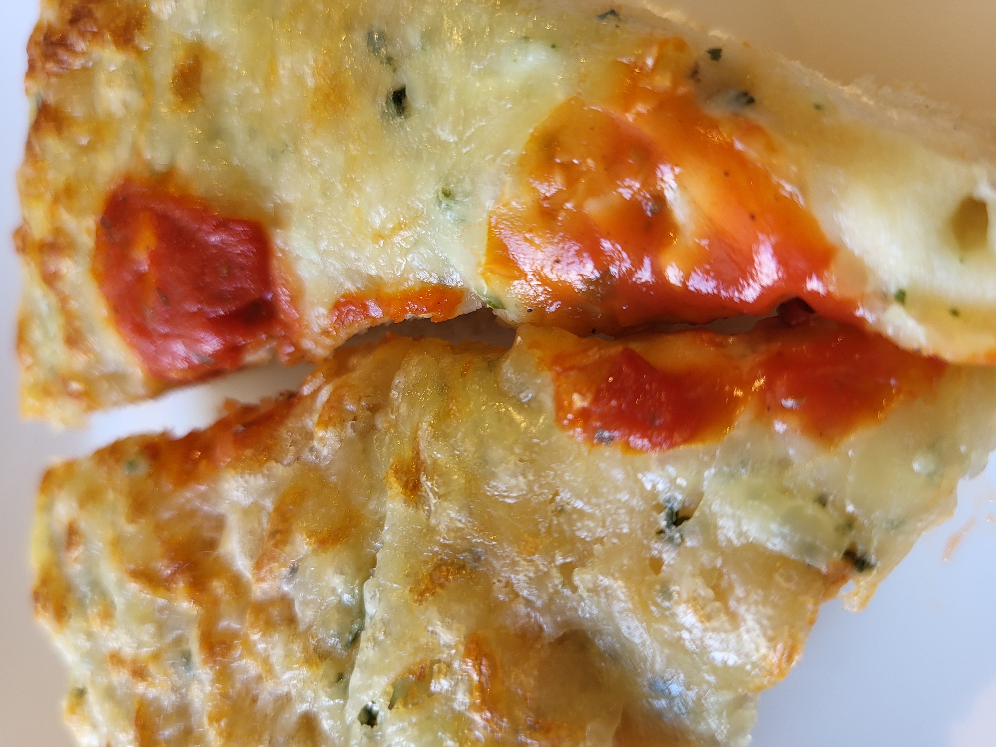 Two slices of pizza are shown in a close-up, top-down view. The pizza has a thick crust that is golden brown and appears to be slightly puffed. The surface is covered in melted cheese, with some areas showing a slight browning and crisping. Red tomato sauce is visible in dollops on top of the cheese, with a glossy sheen. Small green flecks, possibly herbs, are interspersed throughout the cheese. The lighting suggests an indoor setting, possibly a kitchen or dining area, with the pizza resting on a white surface. The location is Unknown, Unknown.