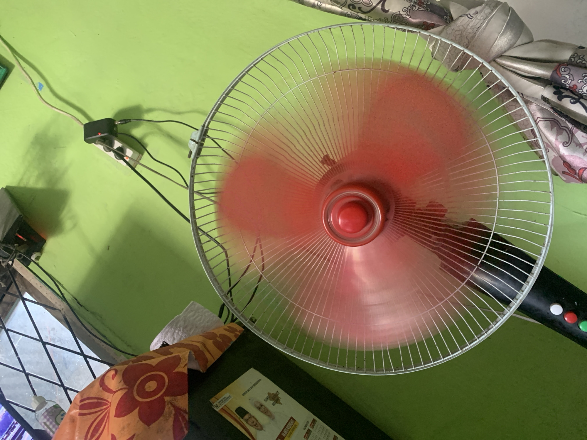 The image shows a red oscillating fan with its blades blurred from movement, suggesting it is on. The fan is positioned in a room with a bright green wall. A window with a diamond-patterned metal grate is visible in the upper left corner. Electrical outlets and cords are present on the green wall. To the left of the fan, a portion of a black object, possibly a piece of furniture or an appliance, is visible. A patterned fabric with floral motifs is draped over this object. A pamphlet or poster with images of two individuals and text is also partially visible on the black object. In the lower part of the image, the base of the fan with three colored buttons (white, red, and green) is seen. A patterned fabric, likely curtains or upholstery, is visible to the right of the fan. The overall scene suggests an indoor setting within a residential or commercial space in Palembang, Indonesia.