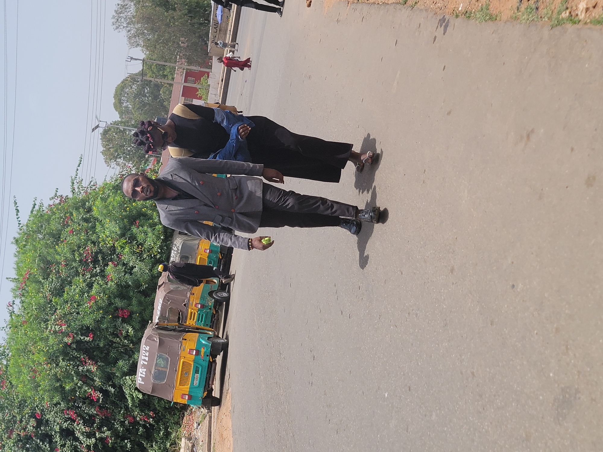 Two individuals walk alongside a row of parked auto rickshaws on an asphalt road, presumably in the city of Jos, Nigeria. The first individual, a male, wears a gray suit and dark shoes, holding a small green fruit in his hand. The second individual, a female, wears a black top, dark pants, and light-colored sandals. They are walking next to a cluster of green leafy plants with red flowers. The auto rickshaws are predominantly yellow and green with the license plate "PTA-7122" visible on one. The weather appears to be sunny and bright. In the background, other people can be seen walking by a building. Electrical wires are also visible in the sky.