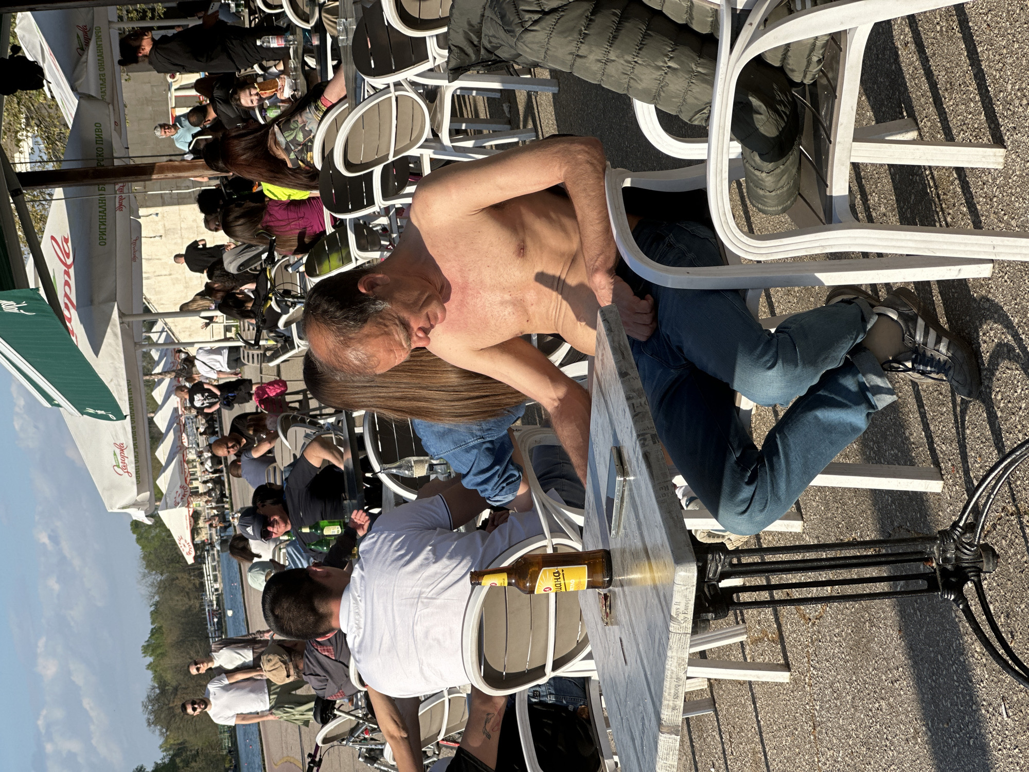 The image captures a busy outdoor scene in Plovdiv, Bulgaria, during daylight. Numerous people are seated at white tables and chairs under "Zagorka" brand umbrellas. One bare-chested man, wearing jeans and sitting on a white chair, dominates the foreground. Next to him, a man in a white shirt is seated. A bottle of "Zagorka" beer rests on their table. The area appears to be a pedestrian zone, with a view of water and trees in the background. Bicycles are also visible among the people.