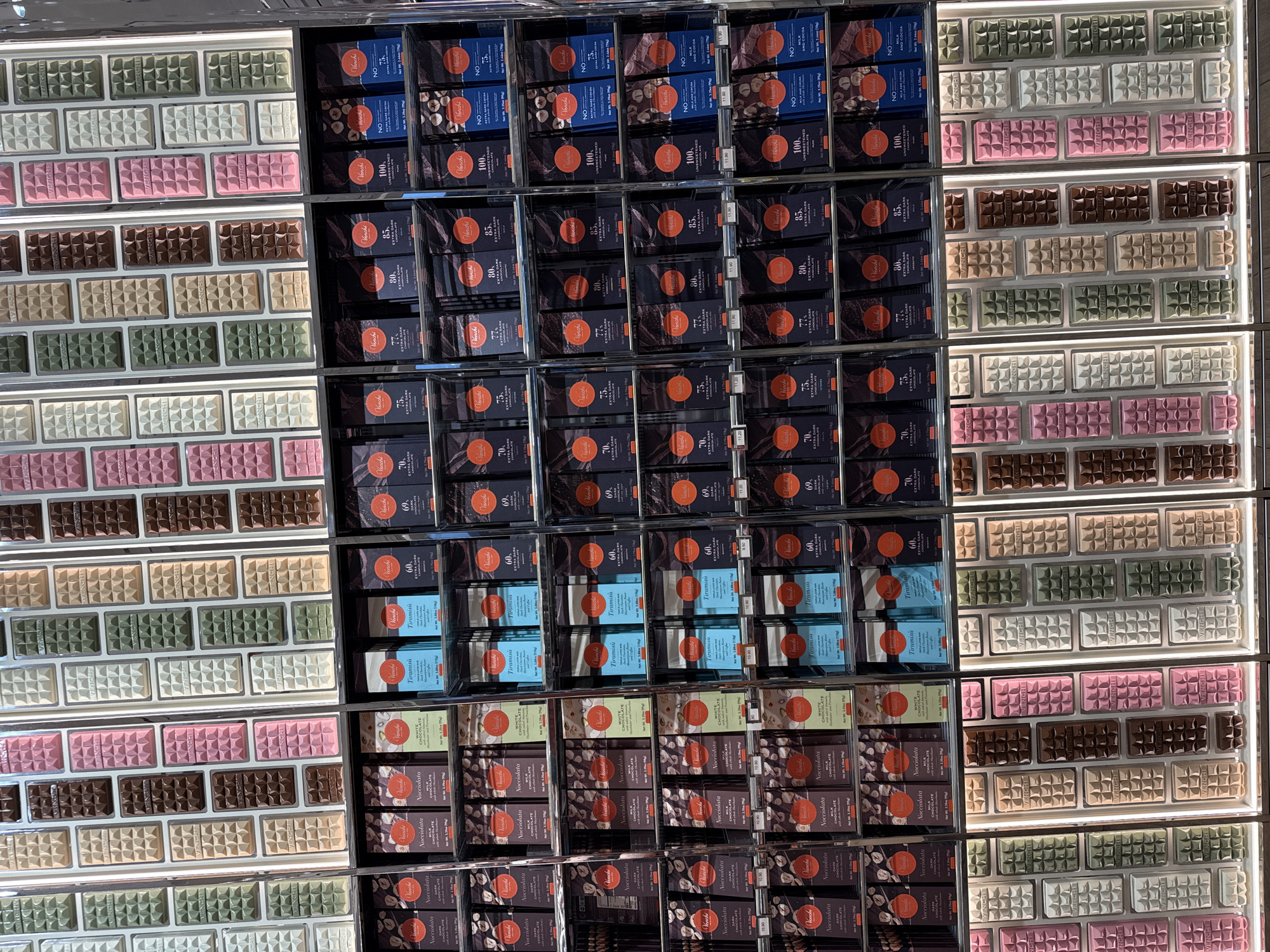 The image shows a retail display of Venchi chocolates in Los Angeles, United States of America. The display features numerous rows of chocolate bars in various colors, including white, pink, brown, and green, with embossed "VENCHI" branding. Below the chocolate bars, there are shelves stocked with packaged Venchi chocolate bars. These packages display different flavors and percentages of cocoa, such as "Nocciolata," "Tiramisu," "Extra Dark Chocolate," and "Unsweetened Chocolate." The lighting in the image suggests an indoor retail environment. There are no people visible in the scene.