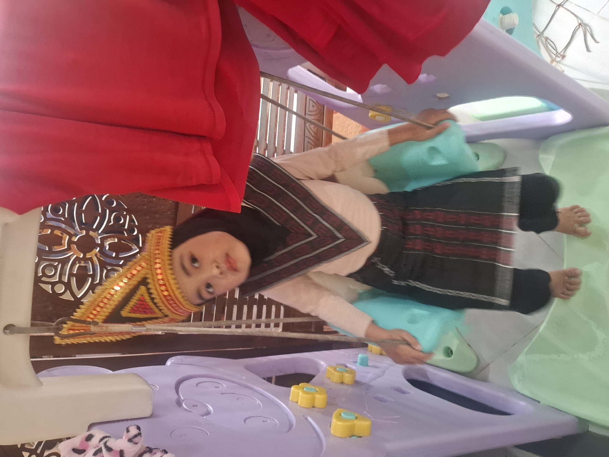 A young girl wearing a traditional gold and red headdress, a black hijab, a light pink long-sleeved shirt, and a black and red patterned skirt stands on a green slide within a multi-colored plastic playground structure. The playground includes purple and light blue elements, with yellow climbing handholds visible on the purple section. The girl is positioned centrally in the frame, looking towards the camera with a neutral expression. Two pieces of red fabric are draped over the top of the playground structure, partially obscuring the background, which appears to be a wooden lattice-like wall with an ornate circular cutout pattern. The scene is indoors, possibly in a residential setting, and the lighting is bright. The image was captured at an eye-level perspective.
