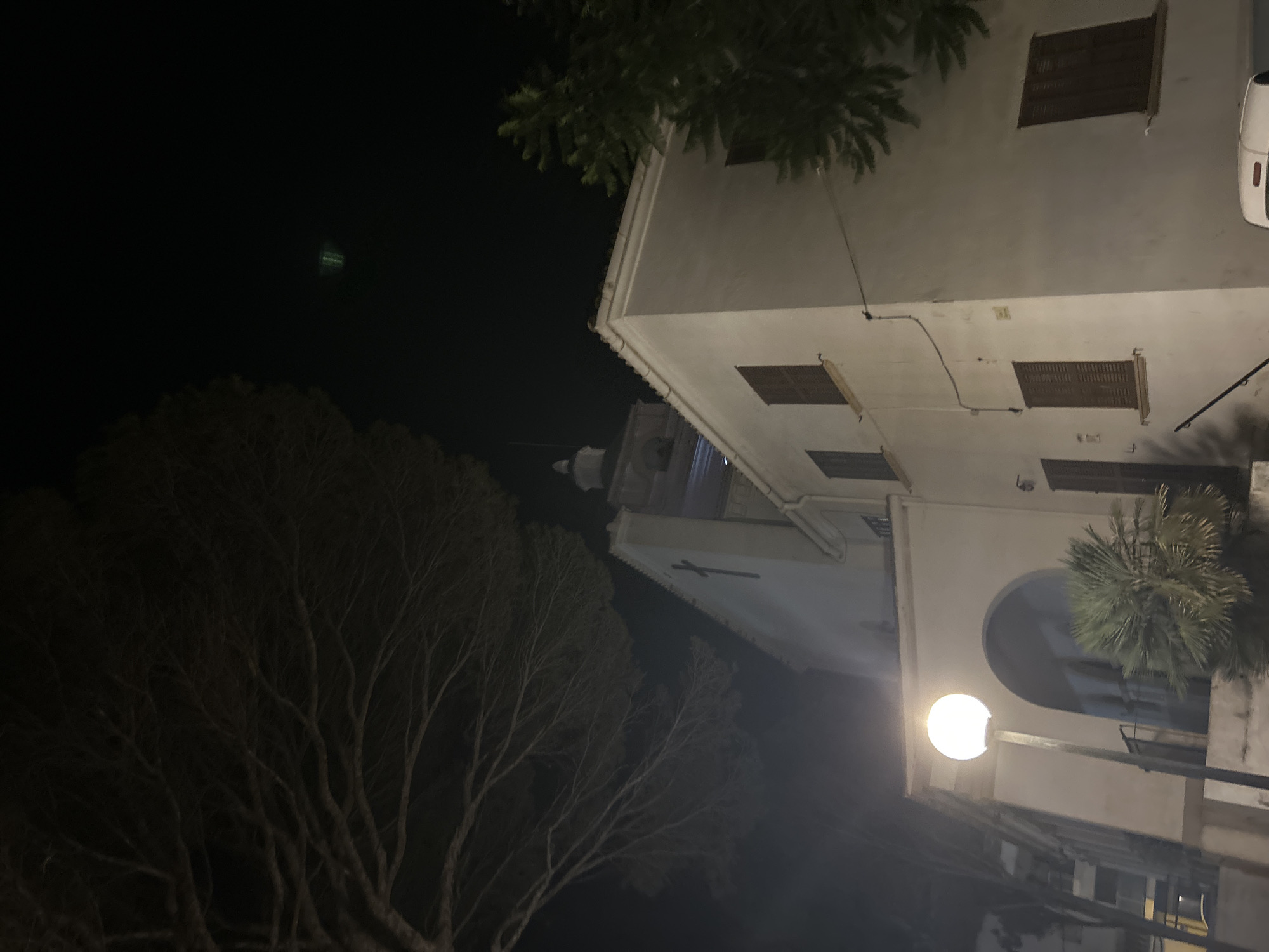 The image shows a building with a white facade and brown shutters, likely in Cala Rajada, Spain. The building appears to be a church or a similar institutional structure, with a prominent cross visible on its side. A bell tower with a dome is partially visible in the background. In the foreground, a street lamp with a glowing orb illuminates the area. Bare tree branches hang over the scene from the left, and a conifer tree is visible on the right. The sky is dark, indicating it is nighttime, and there is some atmospheric haze or fog. The overall scene is dimly lit, with the exception of the street lamp.