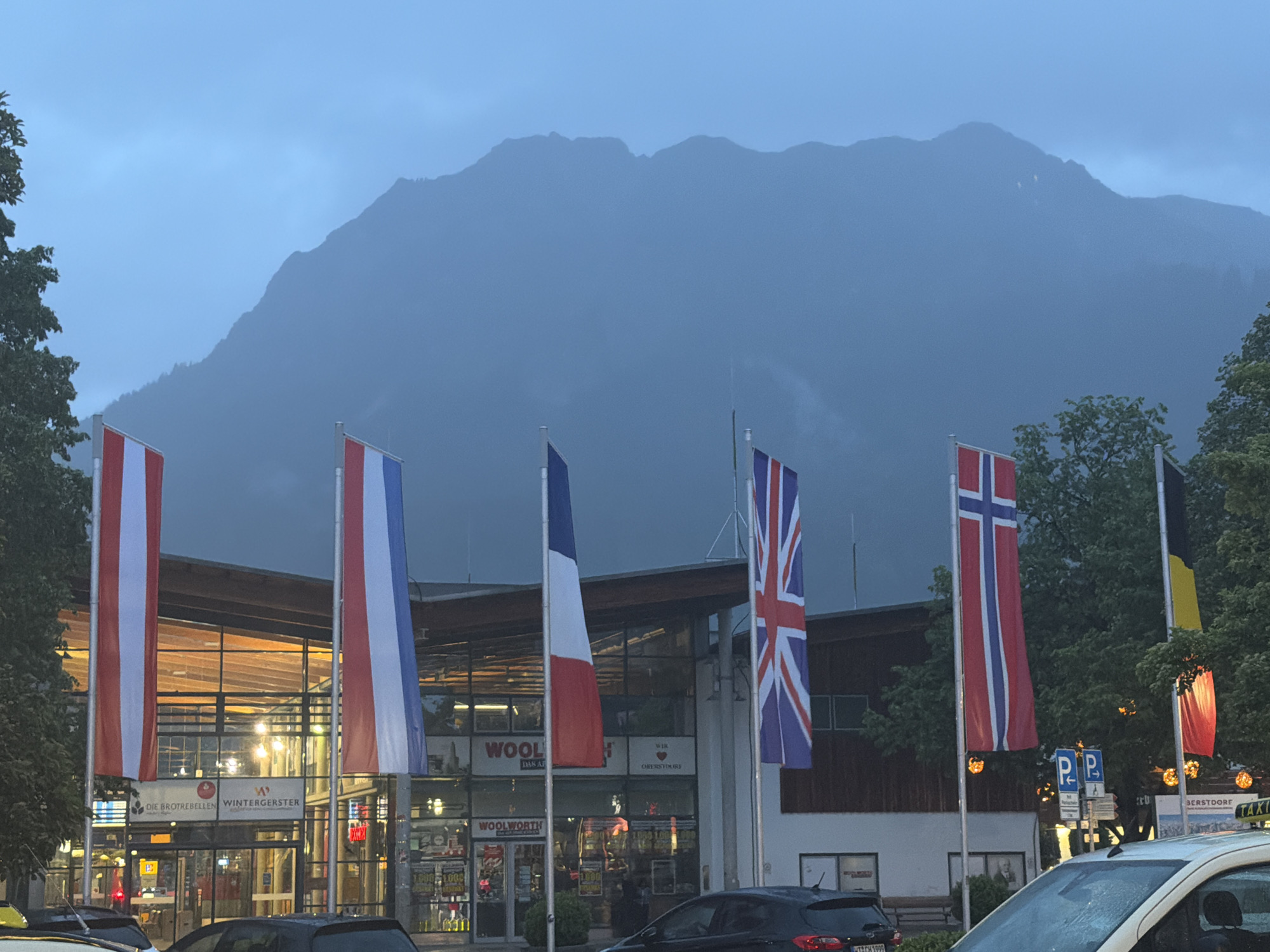 This image captures a street scene in Oberstdorf, Germany, during dusk or early evening. In the foreground, a row of vertical national flags is prominently displayed, from left to right: Austria (red-white-red), France (blue-white-red), the United Kingdom (Union Jack), Norway (red with blue and white cross), and a partially visible flag of Belgium (black-yellow-red). Parts of parked cars, including a white taxi and a dark sedan, are visible at the bottom of the frame, along with a "TAXI" sign and "P P" parking signs.
The midground features a modern building with large glass windows and wooden structural elements, its interior brightly lit, suggesting businesses are open. Visible text on the building includes "DIE BROTREBELLEN", "WINTERGERSTER", and multiple instances of "WOOLWORTH". One sign also proudly states "WIR LIEBEN OBERSTDORF", confirming the location. Trees are interspersed around the building and flags.
In the background, large mountain ranges rise, their silhouettes softly defined and partially obscured by atmospheric haze, mist, or low-hanging clouds. The sky above the mountains is a uniform, dark blue-gray, contributing to the subdued, dusky ambiance of the scene. No people are distinctly visible, and no specific activity or event is taking place beyond the general street scene.