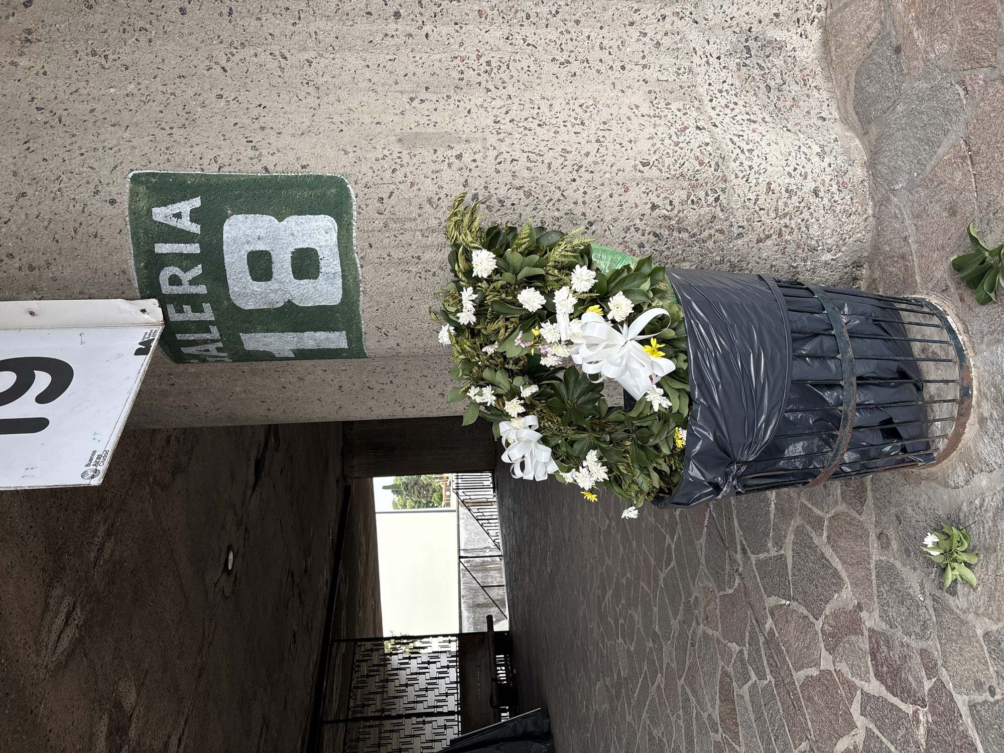 The scene depicts a large floral wreath, primarily composed of white chrysanthemums, green foliage, and white ribbons, discarded within a black plastic bag inside a rusted wire trash receptacle. Several flowers and leaves from the wreath lie on the irregularly paved stone ground. The trash receptacle is positioned against a coarse, light-colored concrete pillar. On this pillar, a green rectangular sign painted in white displays "GALERIA 18." Above it, a partial white sign reveals the number "19" in black and the official "Buenos Aires Ciudad" logo. The environment is an outdoor or semi-covered concrete walkway with a dark ceiling and stone-flagged floor. In the background, an opening reveals a metal bench, a decorative metal grate, and a bright, light-colored structure with trees visible beyond, suggesting an exterior public space. The architectural style and "GALERIA" signage with the "Buenos Aires Ciudad" logo identify the location as one of the entrance "Galerías" of the Estadio Mâs Monumental (River Plate Stadium) in the Belgrano neighborhood of Buenos Aires, Argentina. No people are visible.