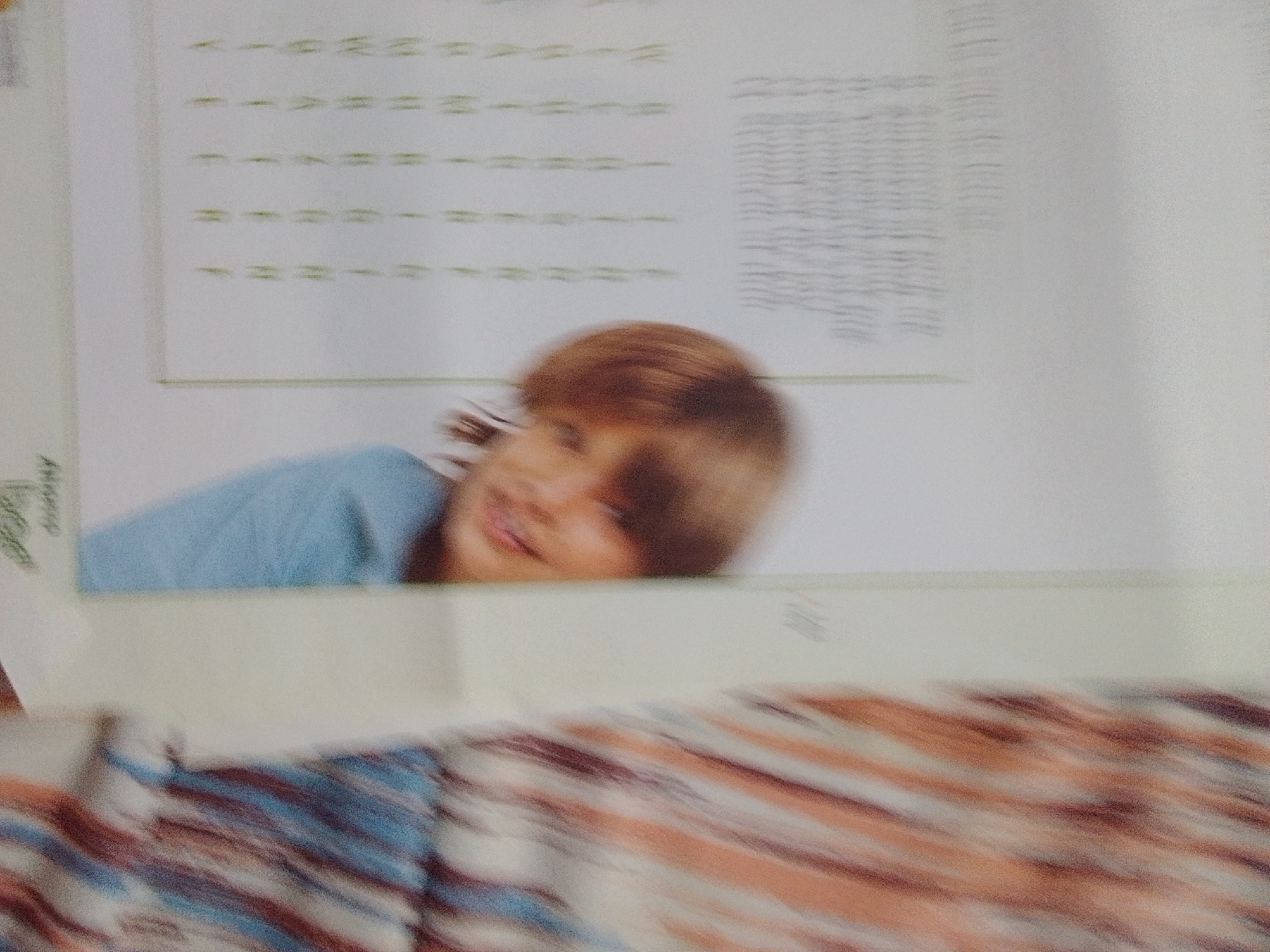 The image shows a close-up of a page with text and a photograph of a child. The text appears to be a list or table, with some lines of characters in green. The photograph shows a young child, with reddish-brown hair, wearing a light blue shirt. The child is looking to the side and smiling. The bottom of the image shows a blurry pattern of diagonal stripes in shades of pink, white, and blue, which appears to be the fabric of clothing or a blanket. There is no information within the image to indicate the location in the city of La Tagua, Venezuela.
