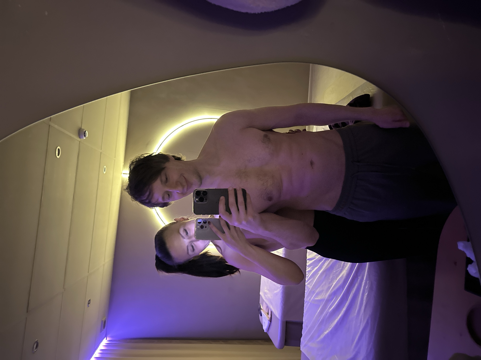 A couple is taking a selfie in a room that appears to be a spa or wellness center. The man is shirtless and wearing grey sweatpants. The woman is wearing a black top. They are both holding up cell phones to take the picture in a curved mirror. The room features white tiled walls, LED lighting, and what appears to be a massage table with white linens. The location is suggested as Saint Petersburg, Russia, but without further evidence, this cannot be confirmed within the image itself.