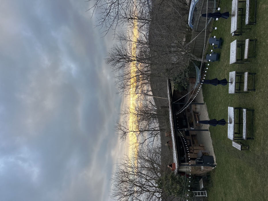 The image depicts an outdoor recreational area, likely a guesthouse or restaurant setting, in Sighnaghi, Georgia, under an overcast sky during what appears to be a cold period, possibly early morning or late afternoon given the light. The upper third of the frame is dominated by heavy grey clouds, with a distinct, bright yellowish-orange band of light visible at the horizon line, suggesting either sunrise or sunset. Bare deciduous trees, characteristic of late autumn or winter, populate the background and midground, with some evergreen foliage visible near structures.
In the midground, a large, rectangular, open-sided covered structure with a dark, snow-dusted sloped roof is present. This structure features brick walls, multiple wooden tables and benches within, and an integrated brick oven or fireplace on its left side. String lights with white bulbs illuminate the interior of this pavilion. Adjacent to the structure, a white step ladder leans against a smaller shed on the far left. A blue hammock is strung between the main covered structure and a tree to its right.
The foreground consists of a green lawn lightly dusted with snow or frost. Four wooden picnic tables, each equipped with attached benches and a folded black patio umbrella, are symmetrically arranged on the grass. These tables also exhibit a light covering of snow. A string of white bulb lights extends diagonally across the lawn from the covered structure towards the right. Two dark-colored Adirondack chairs are positioned on the grass to the right, near a wooden fence. A light-colored vehicle is partially visible behind this fence on the right side of the frame. No individuals are present in the scene.