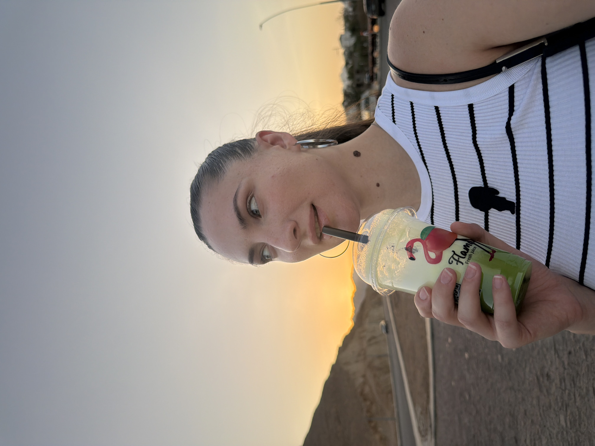 A young woman is shown from the chest up, holding a plastic cup with a green beverage and a black straw. She is wearing a white ribbed tank top with black horizontal stripes and a black graphic of a person. She has a large silver hoop earring in her left ear and a mole on her neck. She is looking to her left, with the sunset in the background casting a warm glow. The scene appears to be outdoors, possibly on a road or a path overlooking a body of water, with mountains in the distance. The sky is a gradient of pale orange and blue, indicating it is either sunrise or sunset. The location is identified as Sharm El Sheikh, Egypt.