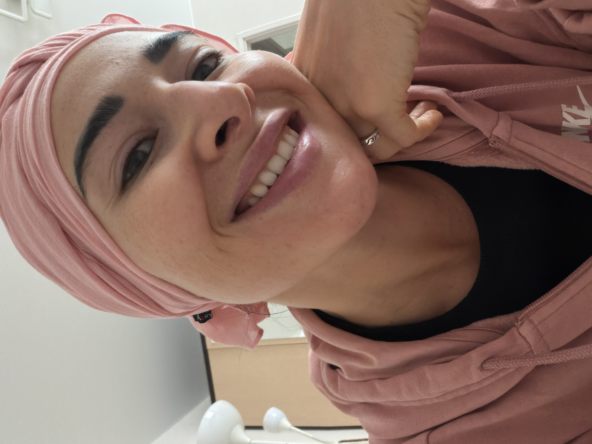 A close-up shot of a woman in a pink headscarf and a pink Nike hoodie over a black top, smiling and resting her chin on her hand, a ring visible on her finger. The location appears to be indoors, possibly a residential or office setting, with a blurry glimpse of a table and wall in the background. The image is rotated, with the woman's head tilted towards the upper left corner. There is no information to determine if the location is Saint Petersburg, Russia.
