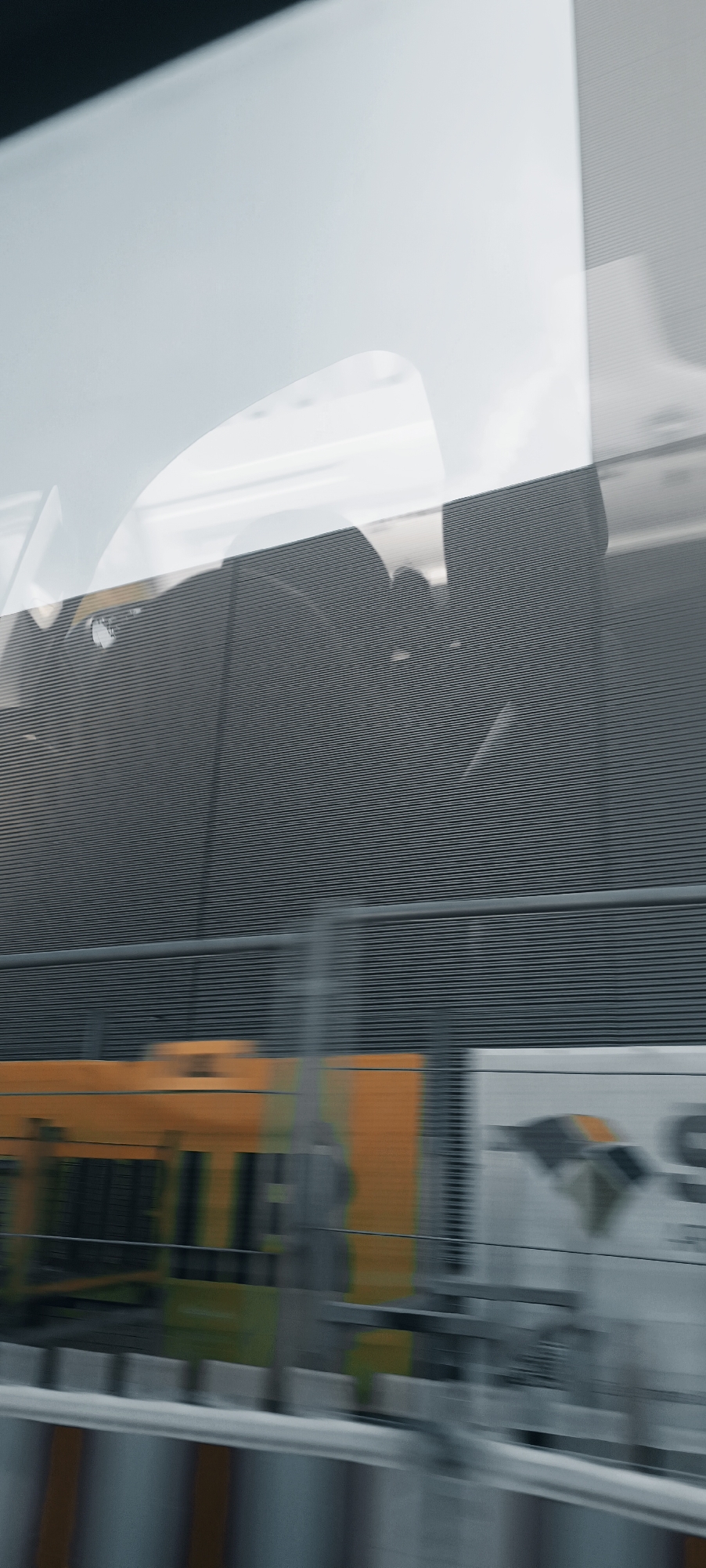The image shows a motion-blurred view of a train traveling through an urban or industrial area. The foreground features a yellow and orange train car with some green accents, suggesting it might be a passenger or freight train. Behind the train, there is a dark gray wall with a horizontal ribbed texture, possibly a sound barrier or the side of a building. A lighter, reflective surface, perhaps a window or another building, is visible above the ribbed wall, showing abstract shapes and reflections. The overall impression is one of speed and transit, with the environment appearing somewhat indistinct due to the motion blur. The location is specified as Winterbach, Germany, implying this is a scene captured while the train was passing through or near the town.