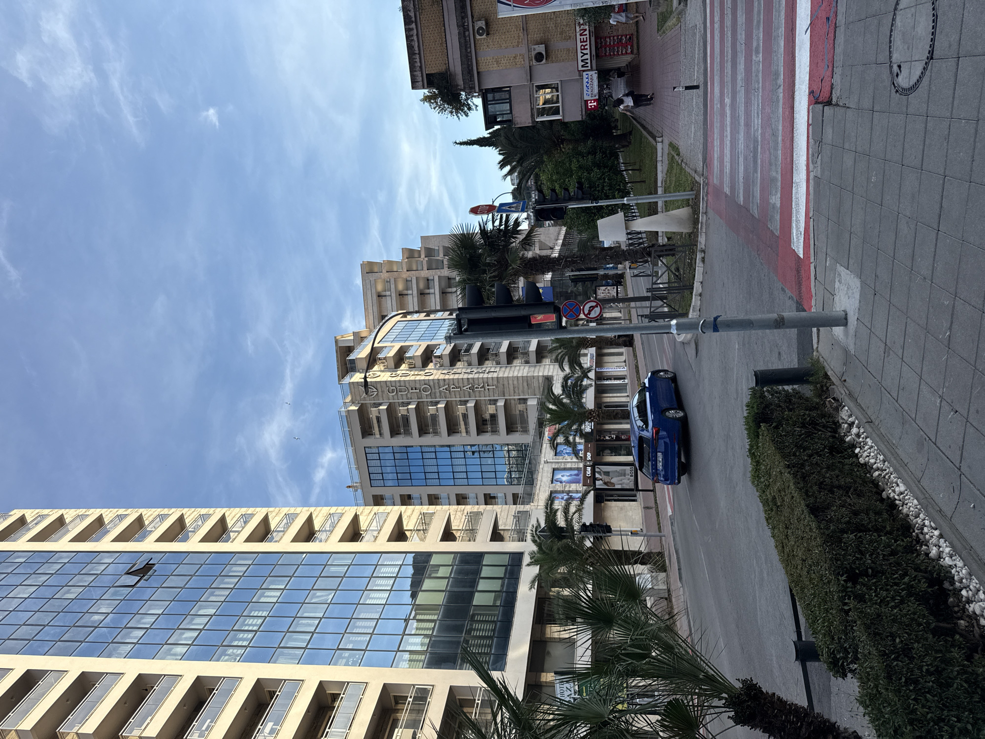 The image depicts a street scene in Budva, Montenegro. Two modern buildings with reflective glass facades and rows of balconies dominate the left and center of the frame. Palm trees are interspersed among the buildings. A blue car is parked on the right side of the road. A pedestrian crossing with red and white stripes is visible further down the road. A building with a "MYREN" sign is on the right, near the crosswalk. There is a sidewalk with gray pavement and a small strip of greenery alongside the road. A few people can be seen in the distance on the sidewalk. The sky is blue with some clouds. Traffic signs are seen near the crosswalk.