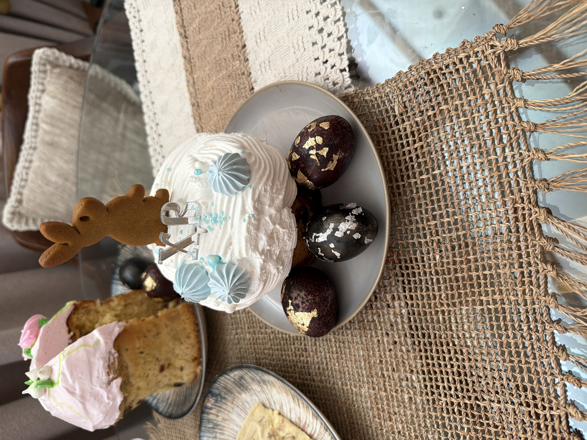 Here is a detailed description of the image:
The image captures a close-up of a table setting likely related to an Easter celebration. The focus is on a light-colored plate containing a decorated Easter cake and several decorated Easter eggs. The cake is round, covered in white frosting, and adorned with light blue meringue swirls, edible glitter, and a gingerbread bunny. The Easter eggs are various shades of brown and black with what appears to be gold or silver leaf accents.
Beside the plate with the cake, there is another plate with a slice of cake topped with pink frosting. The plates are arranged on a light brown woven placemat with a fringe. In the background, there are textures of another light-colored textured table runner or cloth. The table itself appears to be glass. The overall lighting seems natural, indicating an indoor setting.