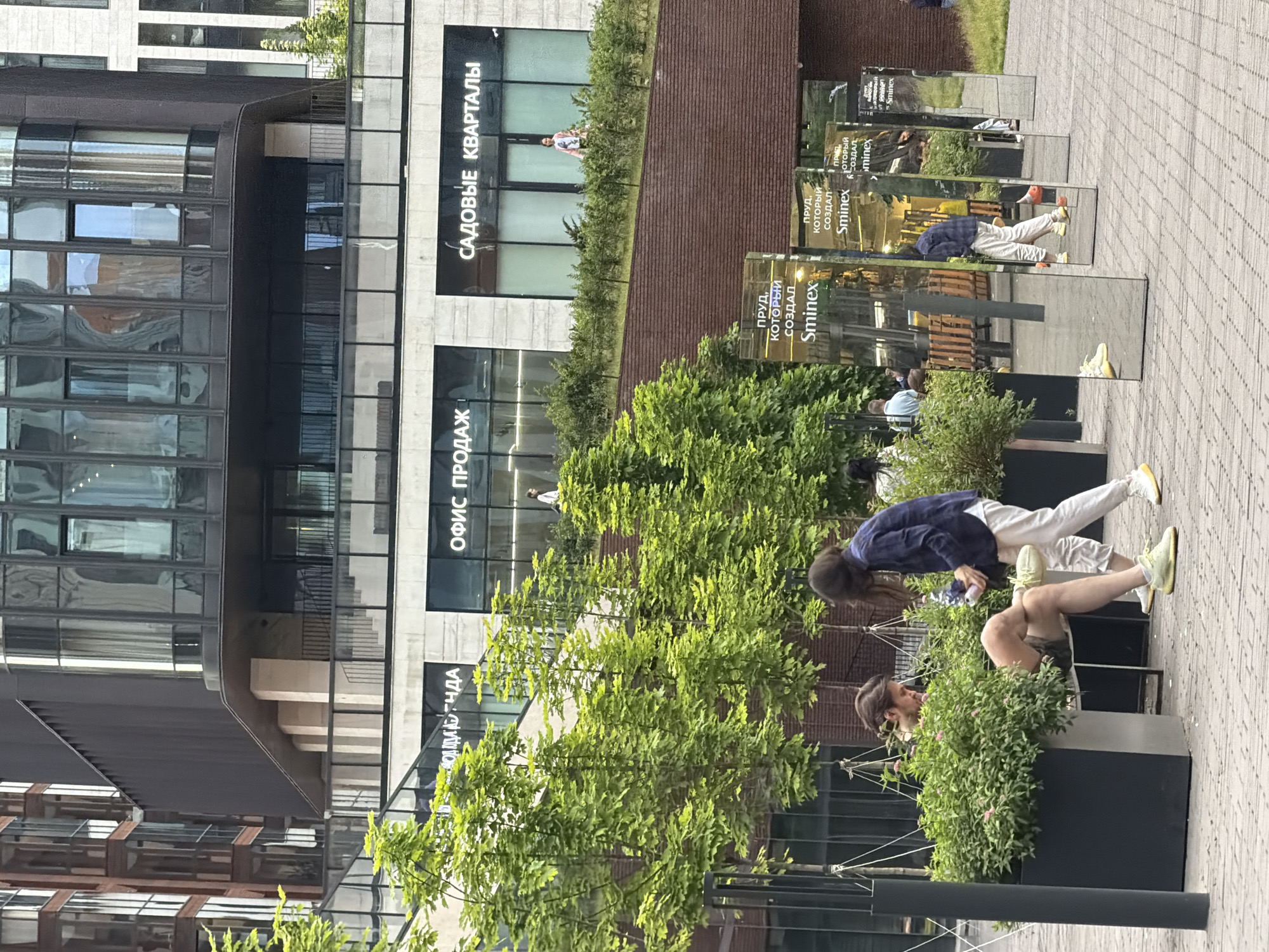 In an outdoor urban setting, a group of people are present. In the foreground, two individuals are interacting, with one seated and the other standing. The seated person is a male with short brown hair, wearing a light-colored t-shirt and shorts, and yellow sneakers. He is sitting in a planter box filled with greenery and small pink flowers, with his legs crossed. The standing person is a female with long brown hair, wearing a dark blue collared shirt and light-colored pants with yellow sneakers. She appears to be holding a white object.
Behind them, a row of tall, rectangular mirrors are placed vertically on the paved ground, reflecting the surrounding environment. Text on the mirrors reads "пруд, который создал Sminex". Further in the background, there are more people visible sitting on benches.
Above, a multi-story modern building complex is visible. The building features dark-colored cladding, large glass windows, and balconies. Signage on the building includes "ОФИС ПРОДАЖ" and "САДОВЫЕ КВАРТАЛИ". Trees with vibrant green foliage are interspersed throughout the scene, adding to the urban landscape. The ground is paved with interlocking bricks.