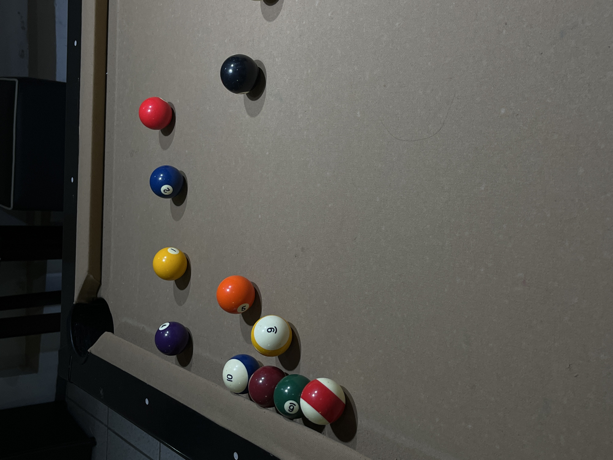 The image shows a billiard table in Unknown, Unknown. The table is covered in a grey cloth. There are twelve billiard balls scattered on the table. The balls are various colors and numbered. The balls are not arranged in a standard rack. The billiard table is located in a dimly lit room. A portion of a black object is visible on the left.