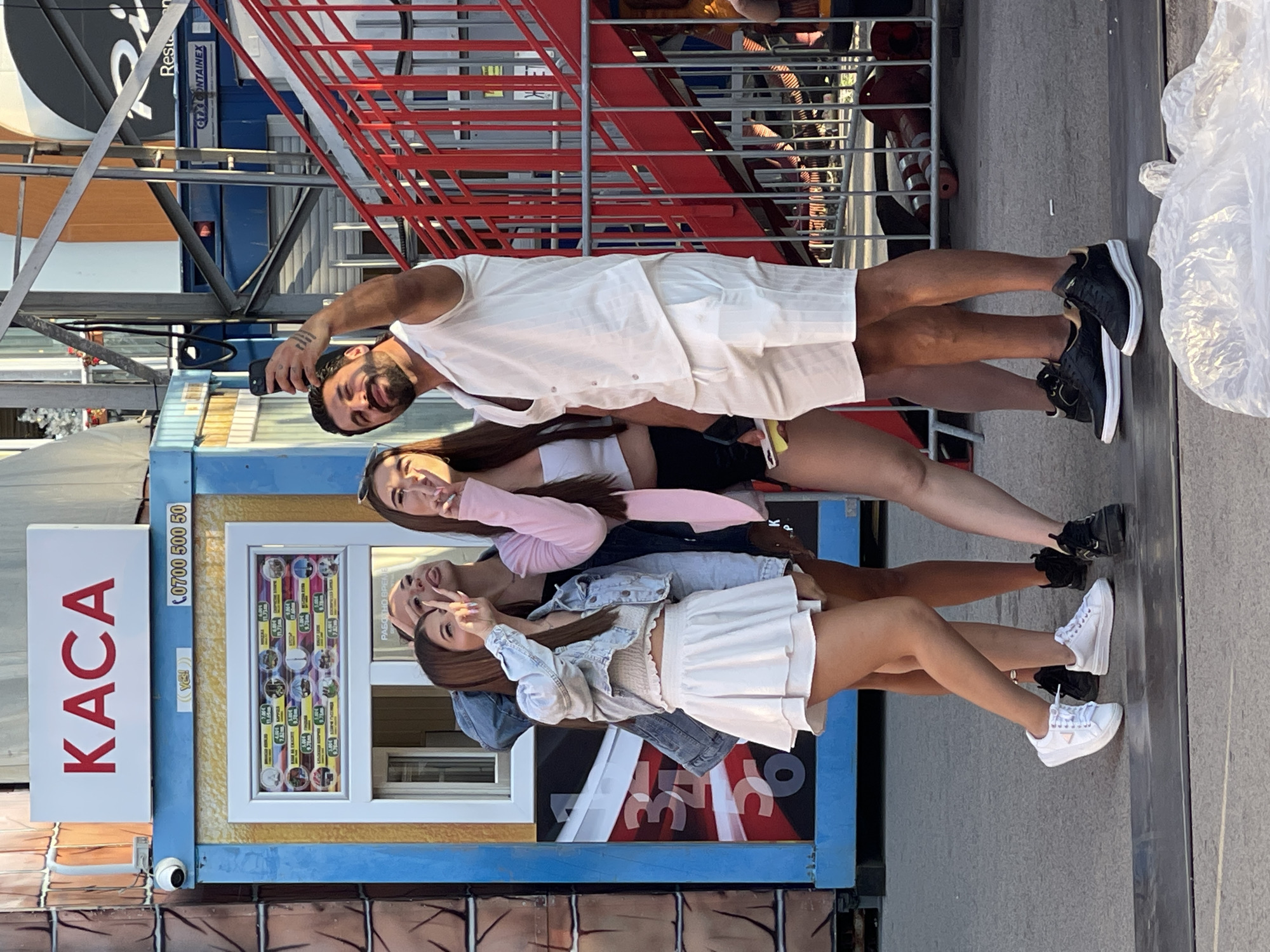 Four individuals, two men and two women, pose for a selfie in front of a ticket booth in the city of Varna, Bulgaria. The booth has "KACA" written in red letters above a window displaying various food items and their prices. To the right of the booth, a red metal staircase with a railing is visible, and beyond that, a fence and some construction materials. The group is dressed casually, with the women wearing crop tops and skirts, and the men in shirts and shorts. The woman on the far left is wearing a denim jacket and white sneakers, while the woman next to her sports a pink blazer, black shorts, and black sneakers. The man on the far right is in a white shirt and shorts, and black sneakers. The man taking the photo is also in a white shirt and shorts. The ground is paved with asphalt, and there is a large piece of white plastic in the foreground. The overall setting appears to be an outdoor entertainment venue or fairground.