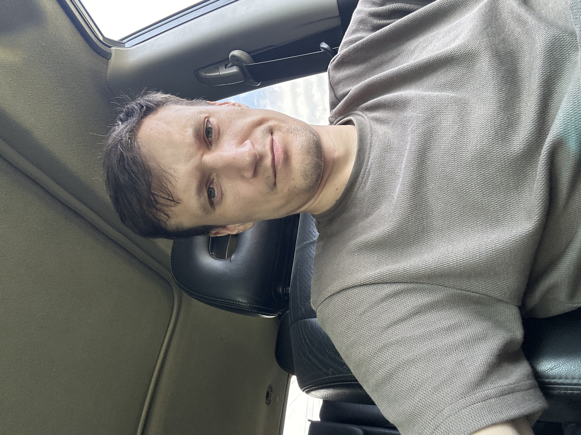 A man with short brown hair and a slight stubble is taking a selfie in the driver's seat of a car. He is wearing a taupe t-shirt. The car interior is visible, including the black leather seats, the sunroof, and the side window. The weather appears to be overcast. The location is unspecified, but the prompt states it is in the city of Makiv, Ukraine.