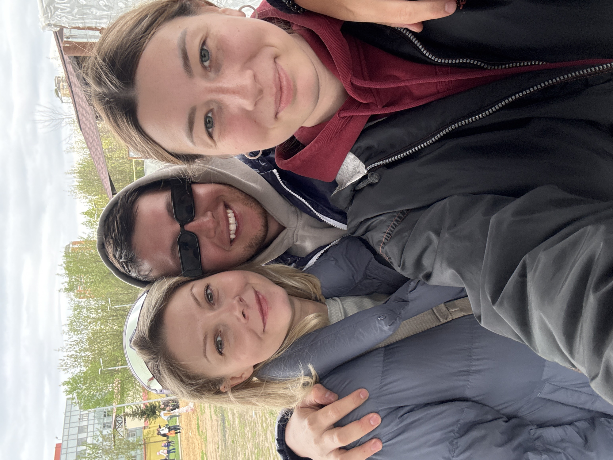 Three people are posing for a selfie outdoors, seemingly in a park or public area. The person at the top has short brown hair and is wearing a black jacket with a red hoodie underneath. The person in the middle is wearing sunglasses and a grey hoodie. The person at the bottom has blonde hair and is wearing a blue jacket, with a hand visible hugging them. In the background, there are trees with green leaves, a building, and some people walking. Given the architecture and general environment, the location is potentially the city of Moscow, Russia.