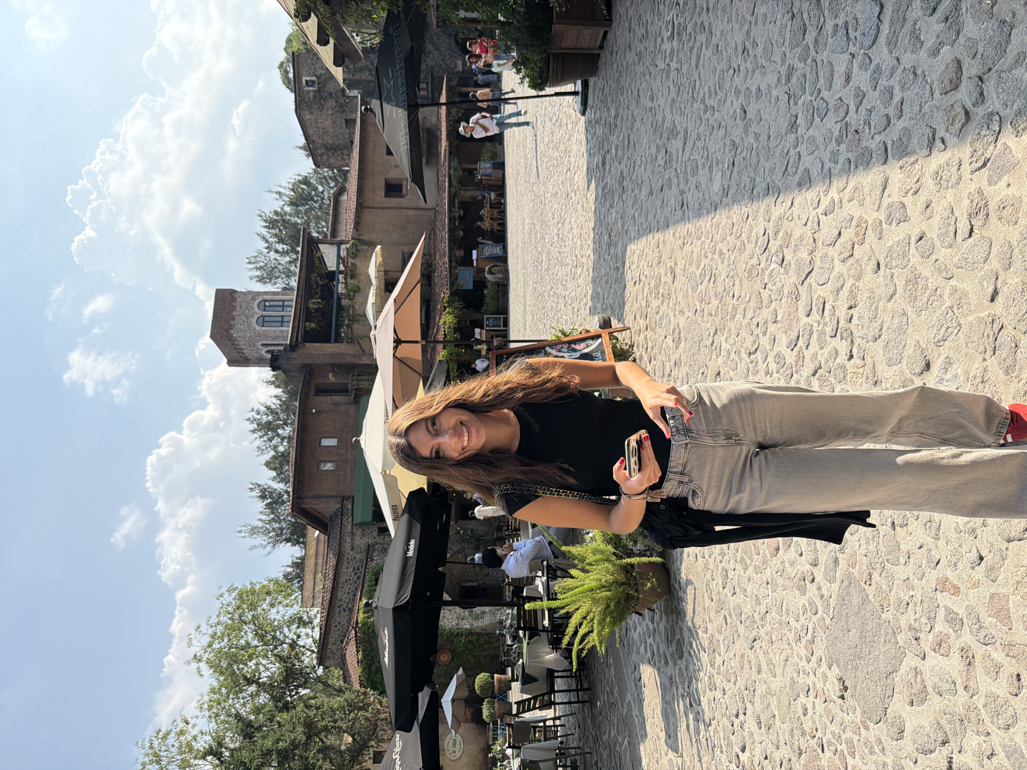 A young woman with long brown hair is smiling and holding a smartphone, seemingly taking a selfie. She is wearing a black T-shirt, light grey jeans, and red shoes. A black sweater is hanging from her shoulder.
The setting appears to be a pedestrian area in Domingo Arenas, Mexico, characterized by cobblestone pavement. There are buildings in the background with a mix of architectural styles, including a stone tower and structures with balconies. Outdoor seating for a restaurant or café is visible, with tables, chairs, and umbrellas. Several people are seated at the tables. The sky is partly cloudy.