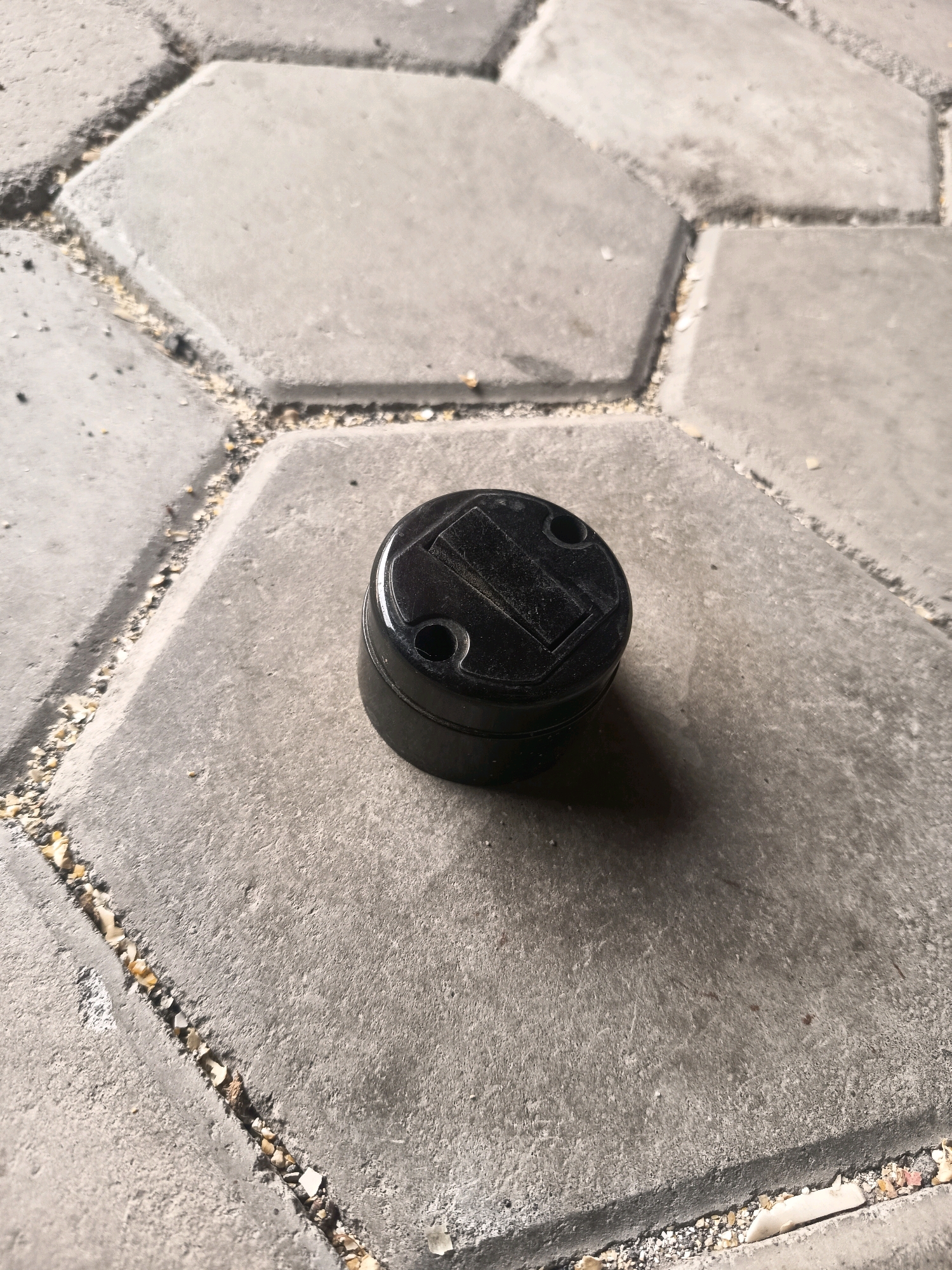 A black, circular, plastic object with a recessed rectangular panel on its top surface rests on a concrete paver pathway. The object has two small circular indentations on its top surface, one on either side of the rectangular panel. The paver pathway is composed of interlocking hexagonal and irregularly shaped concrete pieces with small pebbles and debris filling the gaps between them. The overall scene suggests an outdoor environment, possibly in a public space or residential area in Sidorejo, Indonesia.