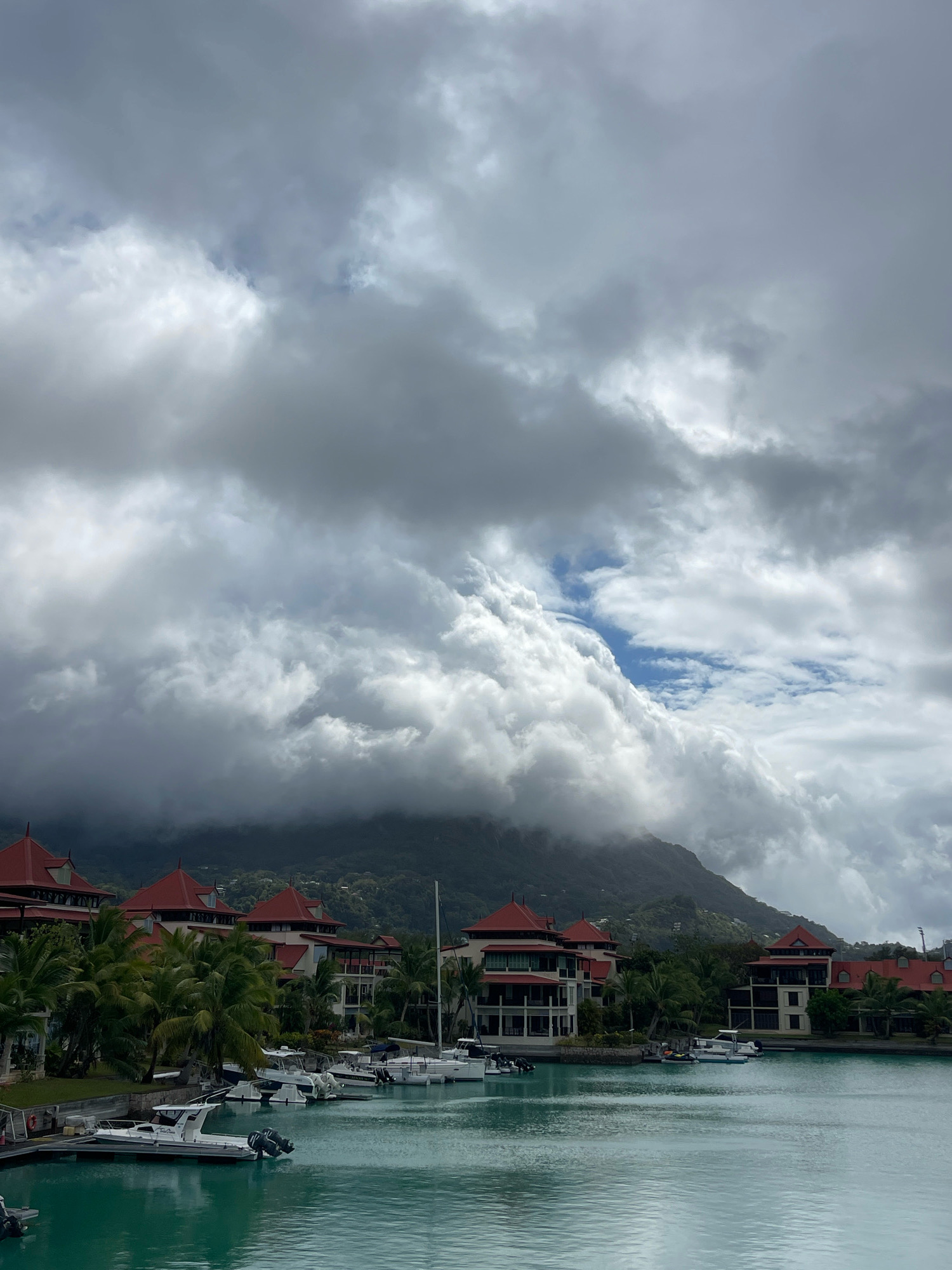 The image depicts a marina with several boats docked in the foreground. Behind the boats, a waterfront development with buildings featuring red roofs and white exteriors is visible, surrounded by palm trees. In the background, a lush, green mountainous landscape is shrouded in dramatic, dark clouds. The water in the marina is a vibrant turquoise. The scene is located in Providence, Seychelles.