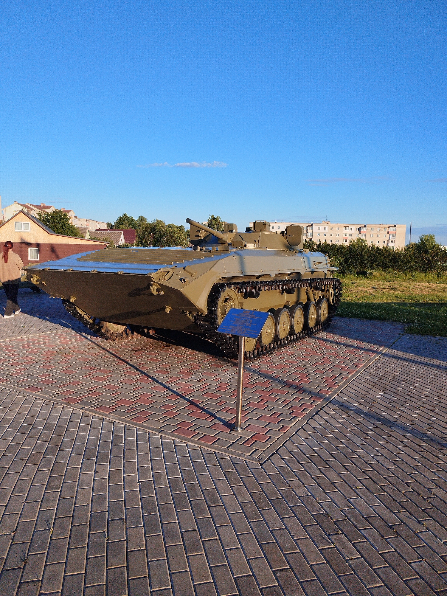 An outdoor, medium shot captures an armored personnel carrier (APC) positioned on a decorative brick paver display. The APC, painted in a matte olive-drab color, is a tracked vehicle with a turret and a large, flat front section that appears designed for amphibious operations. A blue informational sign on a metal pole stands in front of the APC. The APC is situated on a circular section of red and grey brick pavers, which transitions to a wider expanse of grey brick pavers in the foreground. In the background, several residential buildings are visible, along with trees and a clear blue sky. To the left of the APC, a person wearing a light brown hooded jacket and dark pants walks away from the camera. The scene appears to be set in a public space, possibly a memorial or a park, in a city.